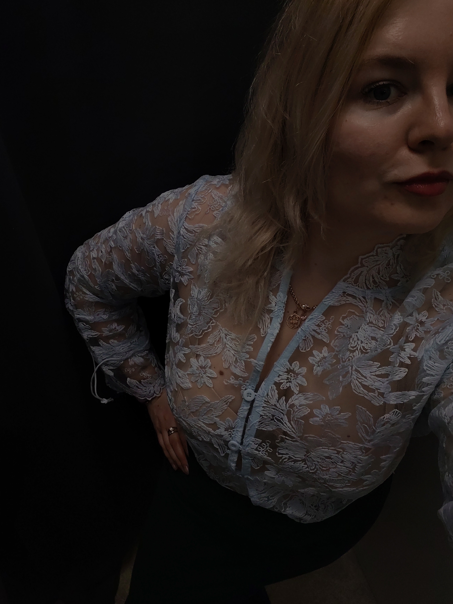 The image is a high-angle, medium shot of a fair-skinned blonde woman wearing a light blue, long-sleeved, floral lace top with buttons and a dark skirt or pants. She is wearing a necklace with a charm. Her lips are red. The background appears to be dark, possibly a dark room. The photograph's location is not identifiable from the provided information.