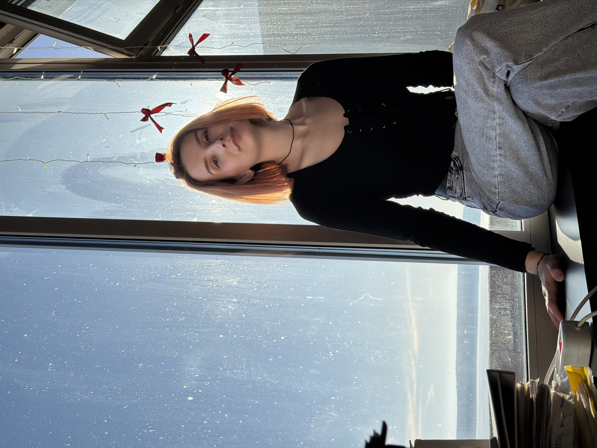 A woman is sitting on a window sill, near a large window with a view, presumably in Chaikovsky, Russia. The window has a metal frame and is decorated with small red ribbons and a string of lights. Outside the window is a bright, potentially sunny scene, possibly overlooking water or a distant cityscape. The woman is wearing a black long-sleeved shirt and gray jeans. Her hair is short and appears to be dyed a reddish-blonde color. The lighting in the room is bright due to the window. The image is rotated 90 degrees clockwise. A desk with items is visible below the window.