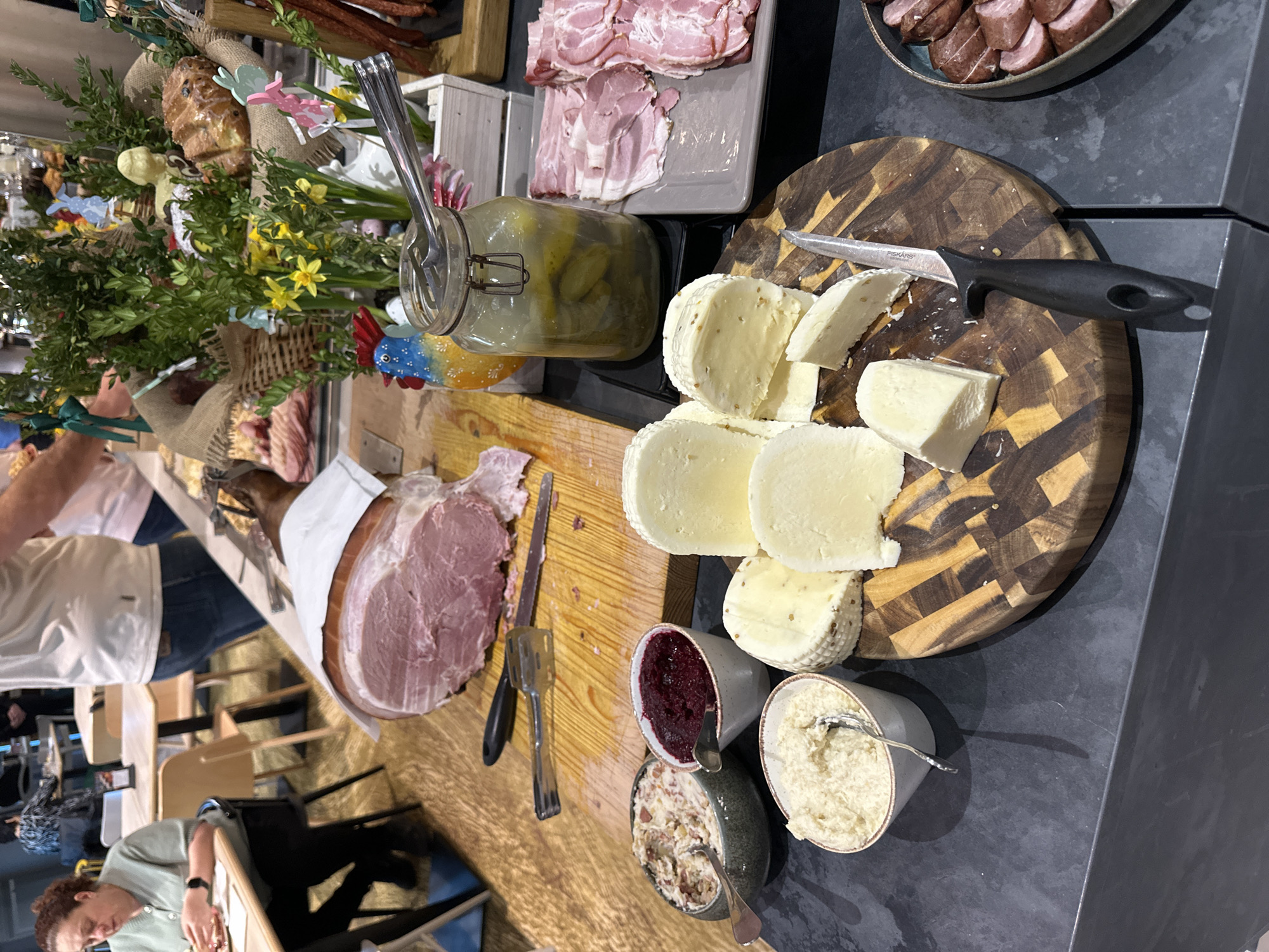 The image shows a buffet table laden with food. Located at the hotel Puro Wrocław Stare Miasto in Wrocław, Poland. The buffet includes various food items such as cheeses served on a wooden cutting board, cold cuts including ham, sausages, and bacon. Additionally, there are jars of pickles, bowls of spreads and ham being cut by a person. There is also a large easter display behind the food. A woman is sitting at a table and may be sleeping. Other people are standing near the buffet.