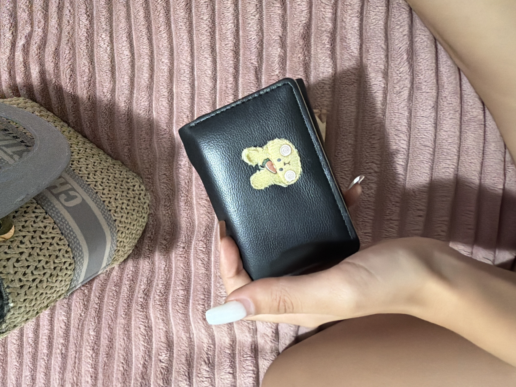 A person's hand with white acrylic nails is holding a small black wallet with a yellow embroidered bunny on it. The wallet is placed on a textured pink surface, possibly a blanket or rug. In the background, a portion of a straw bag with grey straps and lettering is visible. The scene is an indoor setting. There is no information to determine the location in the city of Digah, Azerbaijan.