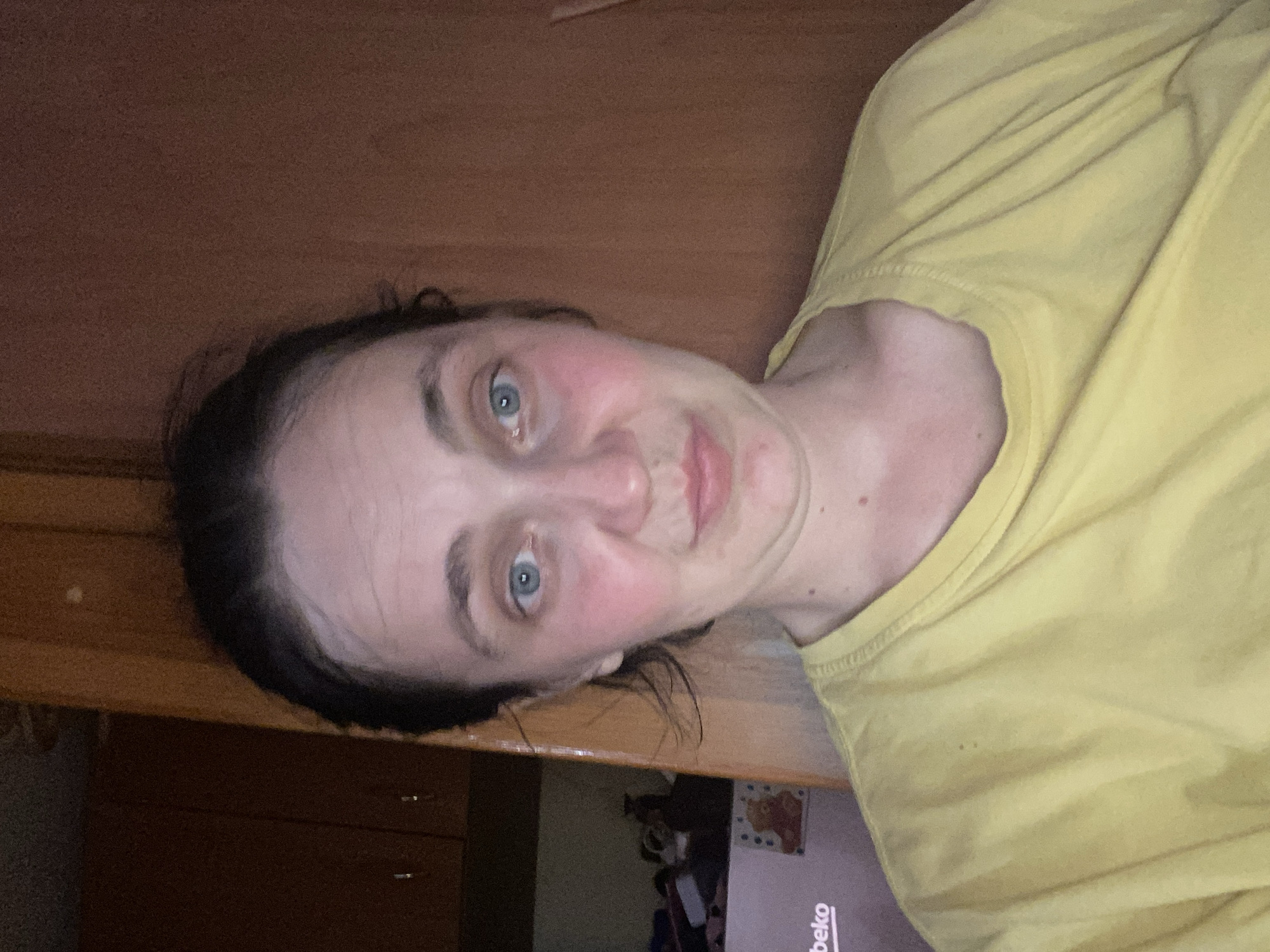 A woman is captured in a close-up, front-facing shot, her face dominating the frame. She has fair skin, tinged with pink across her cheeks, and her blue eyes are wide and direct. Her dark hair is pulled back loosely, with some strands escaping to frame her face. Her expression is neutral, with a slight pout to her lips. She is wearing a plain yellow t-shirt. The background is dimly lit and appears to be an indoor setting, with a wooden surface to her right and what looks like a cabinet or appliance to her left, featuring some faint text and a small, framed image. The overall lighting suggests a low-light environment.