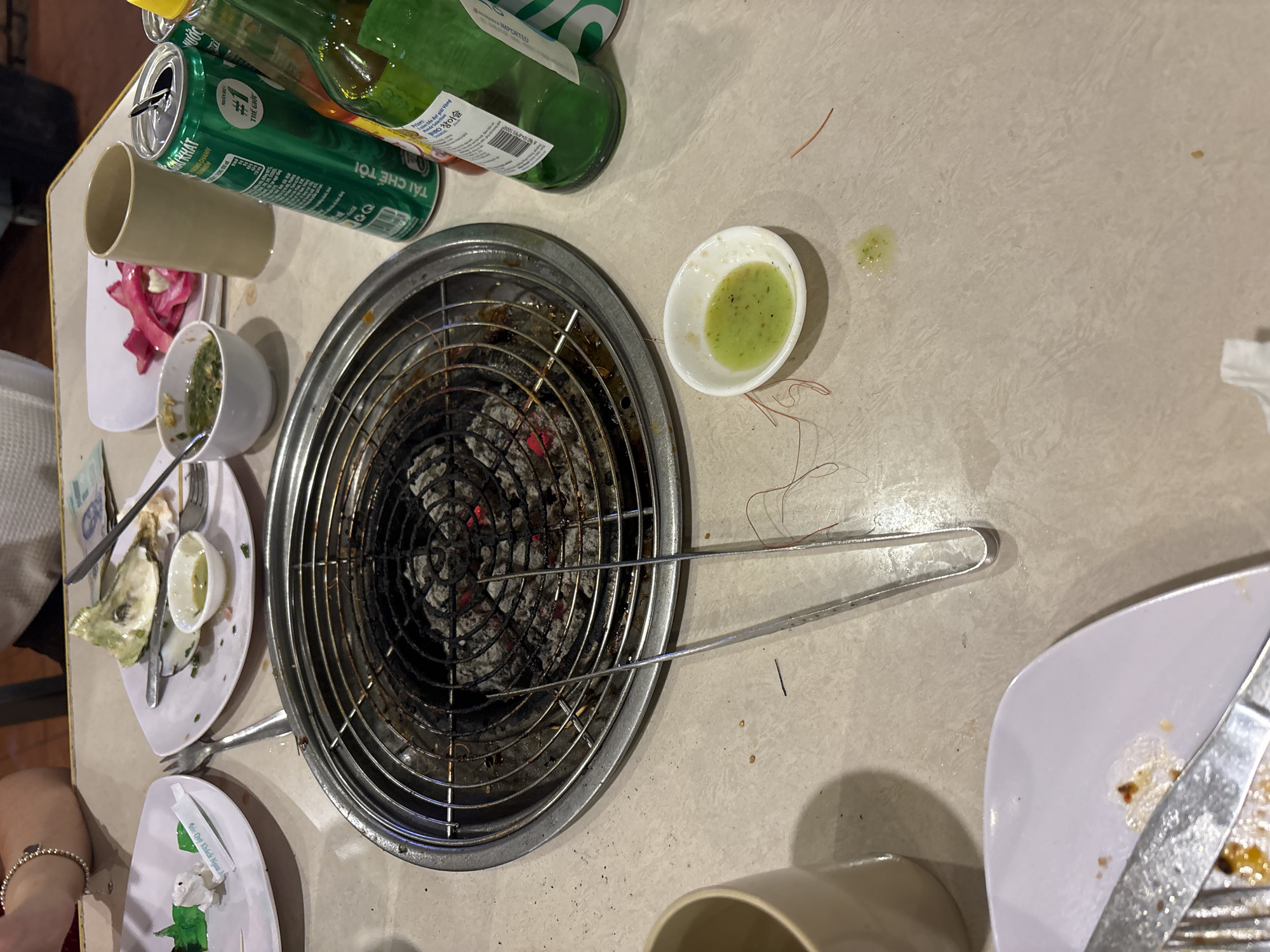 The image presents an overhead view of a dining table, likely in an outdoor restaurant or street food setting in Nha Trang, Vietnam, as indicated by visible text and context. The scene suggests a meal centered around grilling, probably taking place in the evening or at night.
At the center of the table is a small, circular metal charcoal grill with a grate, containing glowing embers and ash, indicating it is or was recently in use. A pair of long metal tongs rests across the grill.
Various dining items are scattered around. On the left, two green beverage cans (one open, one closed) and a green glass bottle are visible. Text on the cans includes "TÁI CHẾ TÔI" (Recycle me) and "#1 THẾ GIỚT" (The world's #1), along with "ONG CHANH". The bottle shows "IMPORTED" and "참이슬" (Chamisul), a Korean soju brand. Below these are a beige plastic cup and several small white plates with meal remnants, including oyster shells and cutlery. One plate holds red/purple leafy vegetables, and another small bowl contains a yellow-green sauce.
To the right, a small white oval dish holds a light green dipping sauce, with some spillage onto the light-colored tabletop. Reddish thread-like debris is also scattered on the table. Part of another white plate with food residue and cutlery is visible on the bottom right. A person's arm with a beaded bracelet is seen on the bottom left, suggesting diners are present. A small white wrapper on a plate reads "Chúc Quý Khách Ngon Miệng" (Wish you a delicious meal), confirming the Vietnamese dining context. The overall impression is of a recently enjoyed, lively meal.