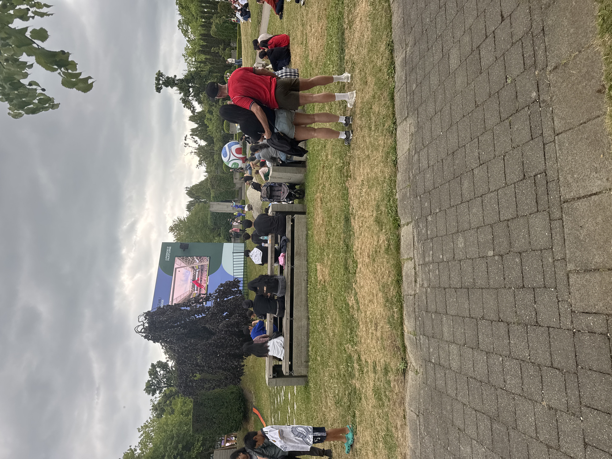 This image depicts an outdoor gathering of people watching a large screen displaying a soccer match. The setting appears to be a park or public space with trees, grass, and paved walkways. In the foreground, a young boy in a soccer jersey stands facing left. Behind him, a group of people are seated at picnic tables. A couple stands in the mid-ground, with the man embracing the woman from behind. In the background, a giant inflatable soccer ball is visible. The screen in the distance shows a soccer game in progress. The overall atmosphere suggests a community event or watch party. Based on the soccer-themed elements and the outdoor park setting, this scene could plausibly be at a location in Vancouver, Canada, that hosts public events, such as a park with a large screen during a sporting event like the World Cup.