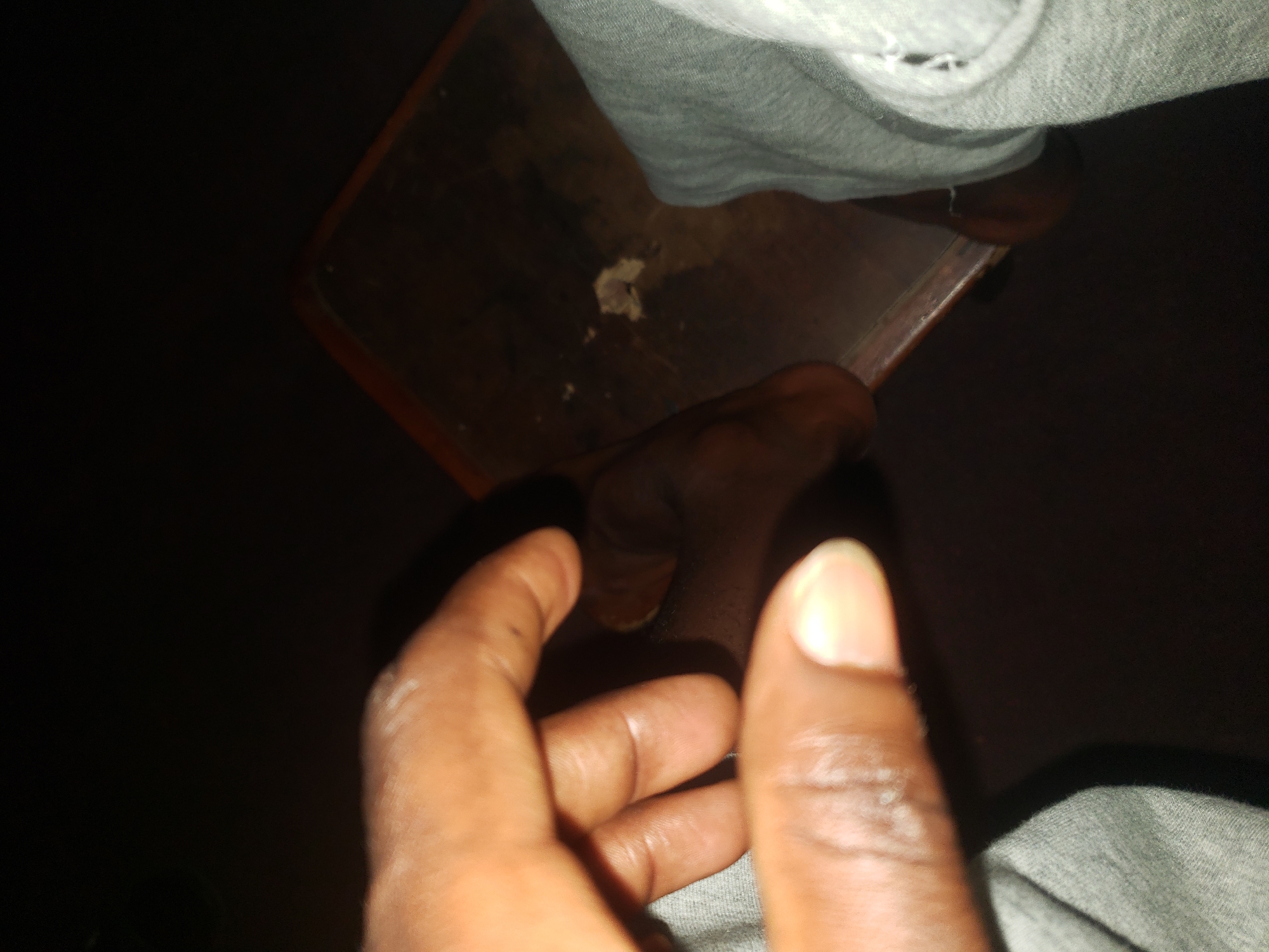 A close-up, low-light, upward-angle shot shows a person's left hand and forearm in the foreground, with skin tones appearing brown. The fingers are partially curled, with the thumb and index finger extended. The hand and arm are resting on a gray, textured fabric, likely clothing. In the background, a dark brown, wooden surface is visible, possibly a table or desk, with a small clump of white material on it. The lighting is dim, creating shadows and obscuring details in the background. The setting is dark and nondescript, with no identifiable landmarks or context to suggest a specific location within a city.