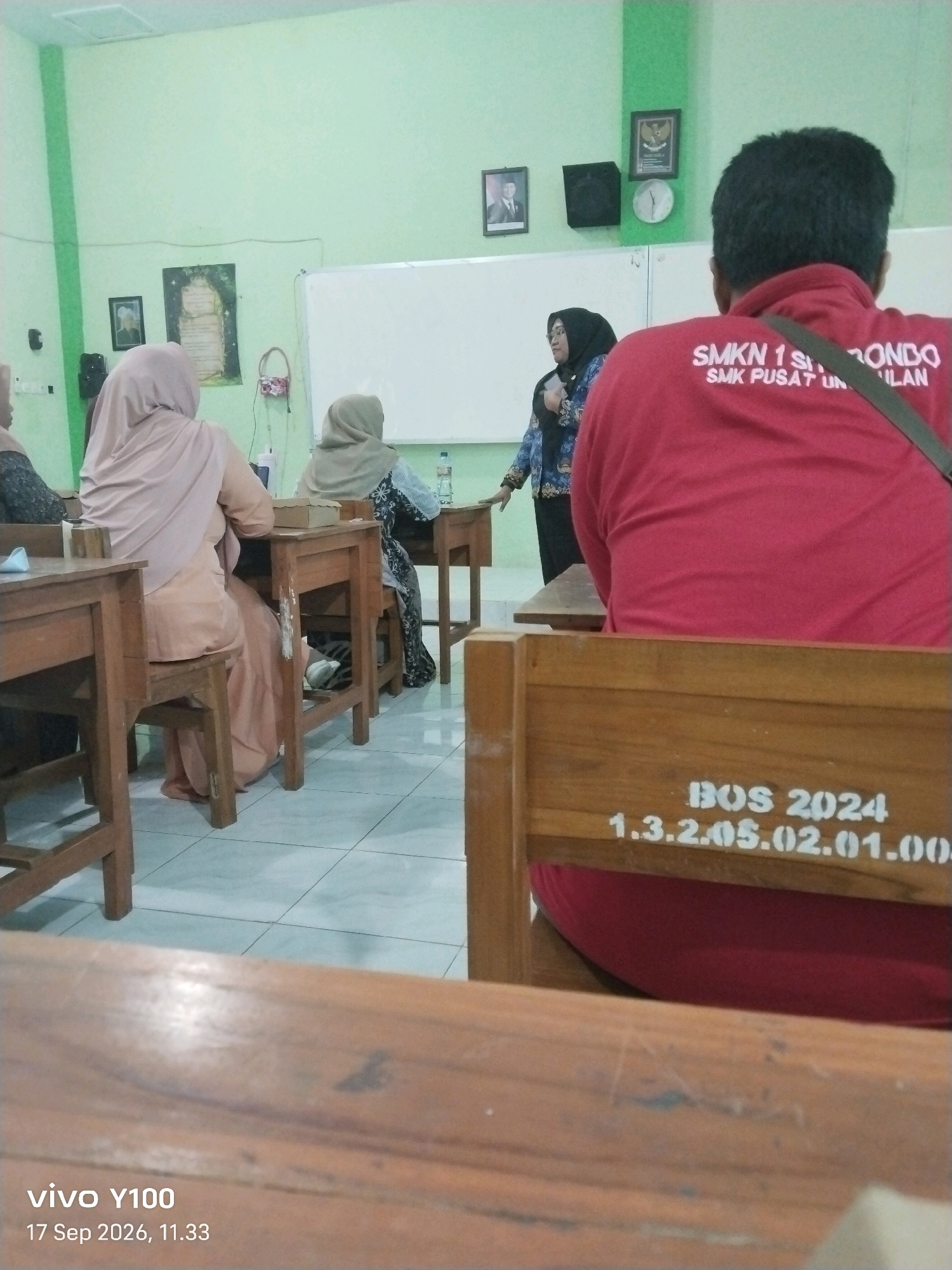 The image depicts an indoor scene, likely a classroom or meeting room, in what appears to be a school setting. The walls are painted a light green, and a whiteboard is visible on the back wall. There are several people present, mostly women wearing hijabs, seated at wooden desks. One woman, standing and facing the seated individuals, appears to be a teacher or presenter, holding an object in her hand. Another person, viewed from behind, is wearing a red shirt with text printed on the back, reading "SMKN 1 S...RONDO SMK PUSAT UN ILAN" and "BOS 2024 1.3.2.05.02.01.004". The overall atmosphere suggests an educational activity is taking place. There are framed pictures and a clock on the wall. The floor is tiled. The date stamp at the bottom left corner indicates "17 Sep 2026, 11.33". The presence of "SMKN 1 S..." strongly suggests this location is within Situbondo, East Java, Indonesia, as SMKN 1 Situbondo is a prominent vocational school in that region.