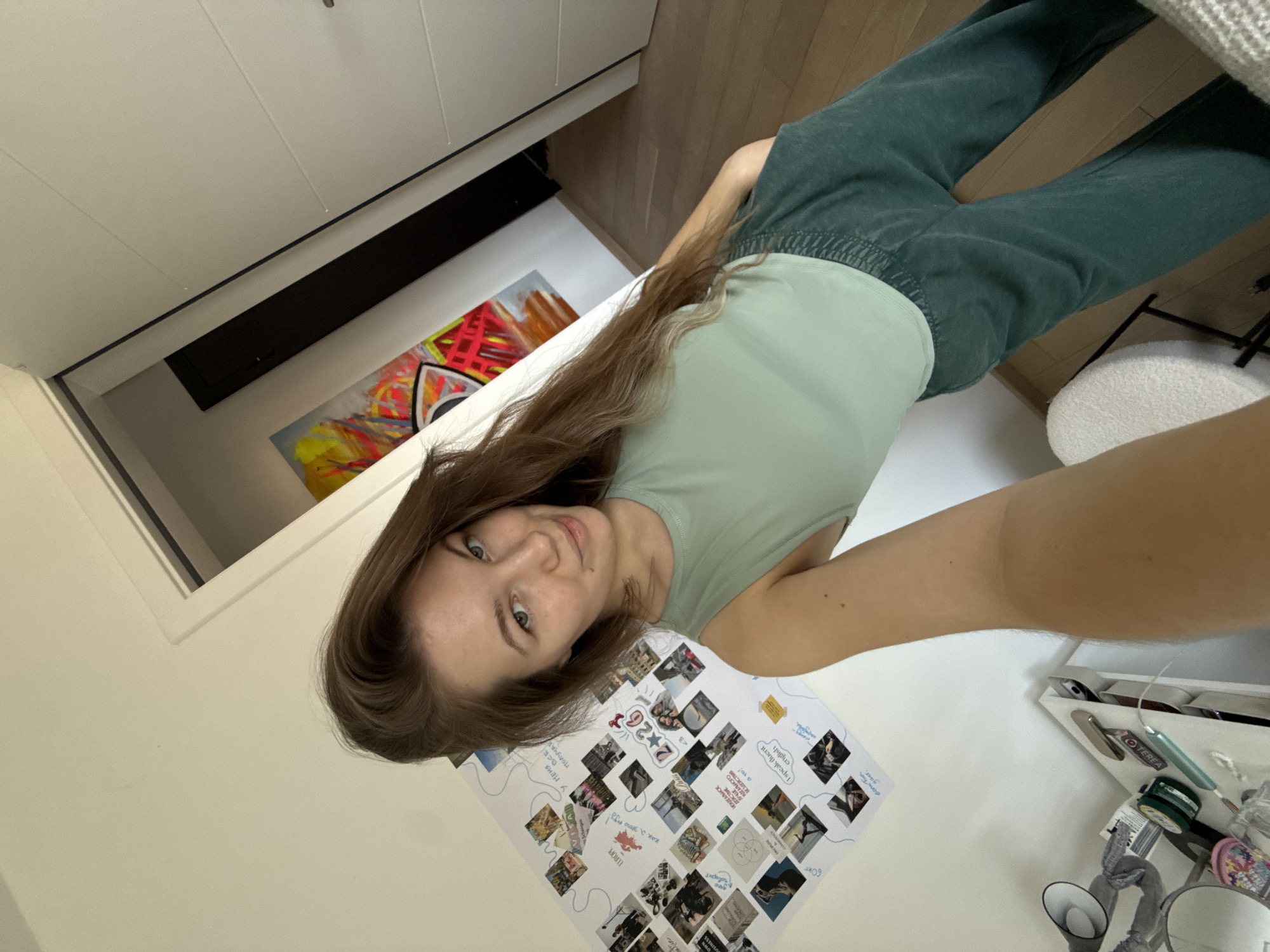 A woman is taking a selfie in what appears to be a room in a residence. She is wearing a light green top and darker green pants. A collage of photos and text is on the wall to her left. The collage includes the following text: "TEREA," "60kg," "I speak fluent english," "Europe," "У МЕНЯ BCE ПОЛУЧАЙ," and "2+26<3". Furniture, including a white cabinet and a wooden surface, are visible in the background. A piece of modern art hangs on the wall. Given the OCR text, it's possible the location is within the broader area of Vozdovac Urban Municipality, Serbia, based on potential language connections.