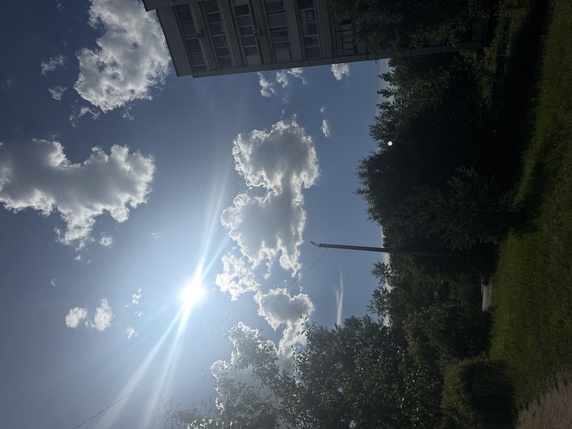 This media file captures a bright, sunny day in Chaikovsky, Russia. The scene is dominated by a clear blue sky filled with fluffy white clouds, illuminated by the sun's intense glare. The sun's rays create a strong lens flare effect, making it difficult to discern fine details in its immediate vicinity.
In the upper left portion of the frame, the corner of a multi-story building is visible, its facade featuring rows of windows. To the right, a dense line of lush green trees forms a natural border. The trees appear to be in full leaf, suggesting a summer or late spring setting. In the foreground, to the lower right, a patch of vibrant green grass stretches outwards, and a dirt path runs alongside it.
A utility pole with a single wire stretching across the frame adds a man-made element to the otherwise natural landscape. The overall impression is one of a tranquil outdoor environment, bathed in the warmth of a sunny day. There are no people or specific activities depicted, and no visible text. The lighting and clear skies suggest it is midday.