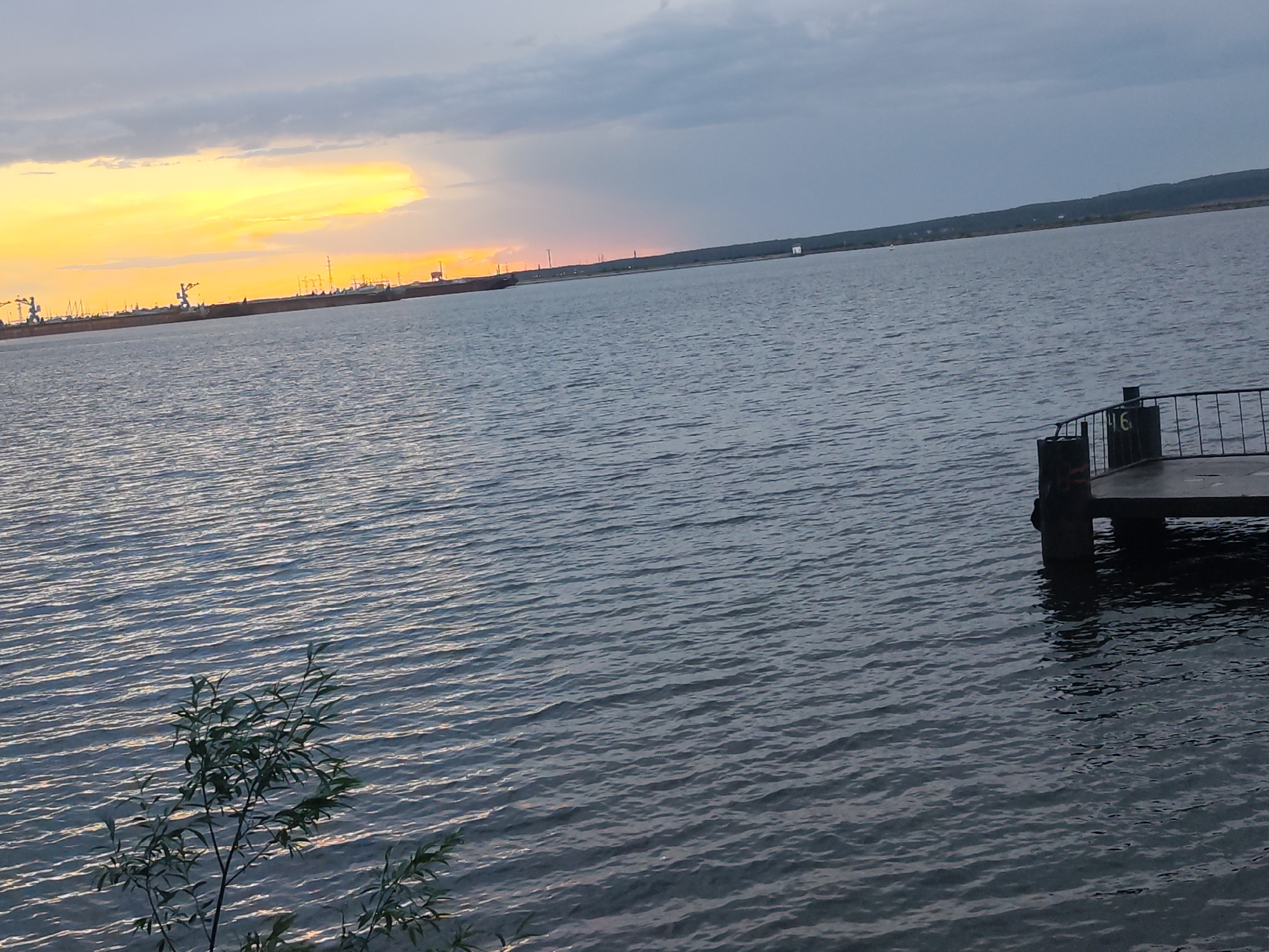 The image captures a body of water with a pier on the right side. The pier has metal railings and the number 16 on one of its supports. In the distance, there are structures that appear to be part of a port or industrial area, with cranes and possibly a large ship. The sky is a mix of dark clouds and a bright, yellow sunset. A small plant is visible in the lower left corner. The location appears to be near a port or harbor, possibly in a city, but further information about the precise location in the city of Unknown, Unknown is unavailable. The water has small ripples across its surface, suggesting a slight breeze.