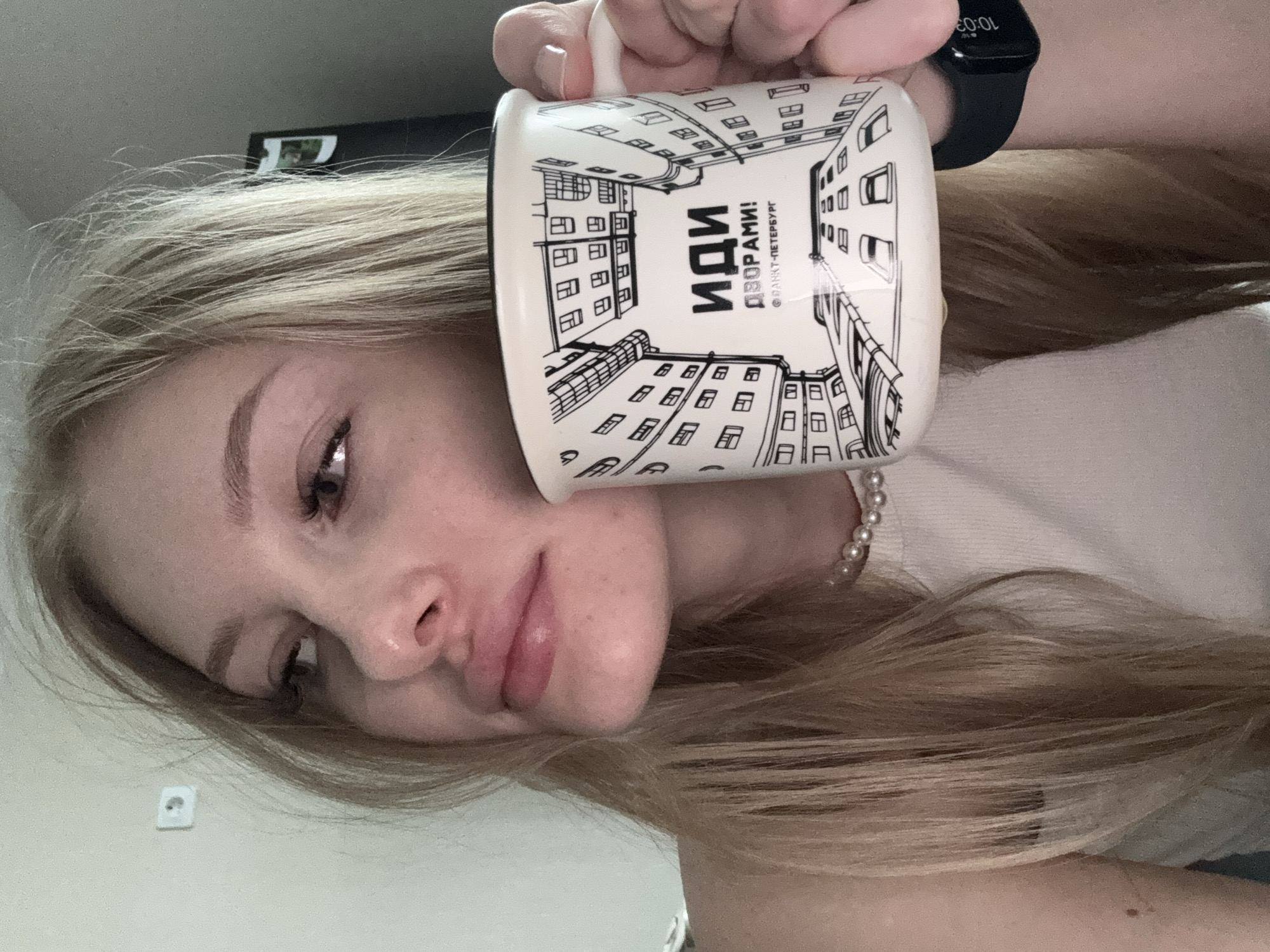 A young woman with long, straight, light blonde hair is depicted in a close-up, chest-up indoor shot, looking towards the viewer's left. She has light skin, full lips slightly parted, and long eyelashes. A pearl necklace is visible around her neck, above a light-colored tank top. She holds a white enamel-style mug with a black rim and handle in her right hand. The mug is decorated with black line drawings of buildings viewed from below, creating a perspective of narrow urban passages. Text on the mug reads "ИДИ ДВОРАМИ!" (GO THROUGH THE COURTYARDS!) and "@РАНКТ-ПЕТЕРБУРГ", a reference to Saint Petersburg, Russia. A black digital smartwatch displaying "10:03" is visible on her left wrist. The background features a plain, light-colored wall with a white electrical outlet mounted on it, and a dark, indistinct object, possibly a refrigerator or cabinet, is partially visible in the upper right. The image contains no visual elements to specify a location in Perm, Russia; the only city referenced is Saint Petersburg.