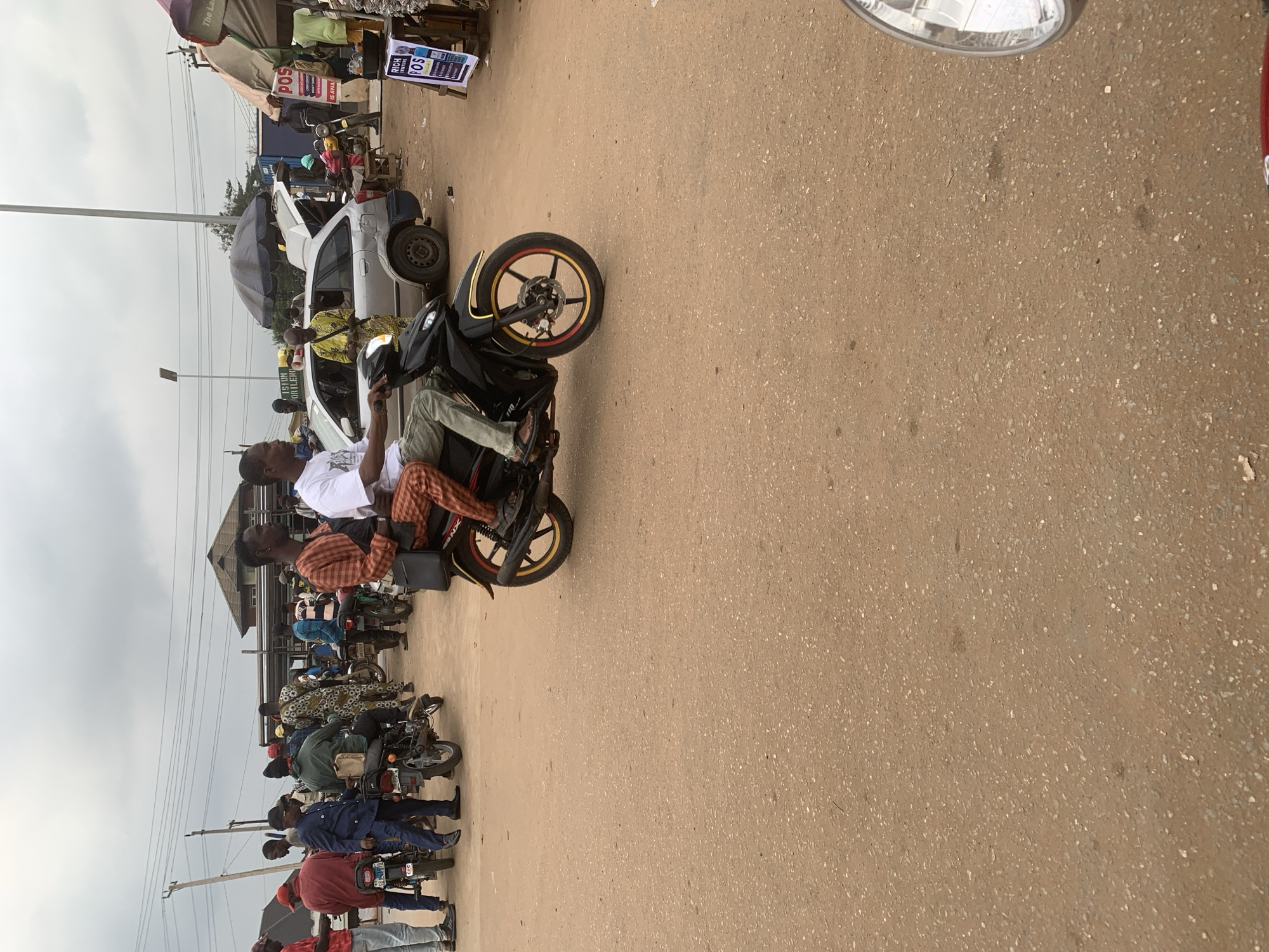 In Shagamu, Nigeria, a bustling street scene unfolds under an overcast sky. The foreground is dominated by a dusty, reddish-brown dirt road. In the middle ground, two men ride a black motorcycle. The driver, wearing a white t-shirt and light-colored pants, navigates the road. The passenger, dressed in a striped orange and brown outfit, is seated behind him, wearing a black bag across his chest.
Further back, a silver hatchback car is parked with its trunk open. Several individuals are gathered around the car and in the surrounding area. To the left, a group of people stand near motorcycles, some of them dressed in casual clothing, others in more traditional attire with colorful patterns. A man in a blue suit and cap stands near a motorcycle.
To the right, market stalls are set up under umbrellas. A sign for "RICH VENTURES POS" is prominently displayed on a wooden stand. Various goods are visible on shelves and tables within the stalls, suggesting a marketplace environment. Overhead, power lines stretch across the sky, and a tall utility pole stands near the left side of the frame. The overall atmosphere is one of everyday activity and commerce.