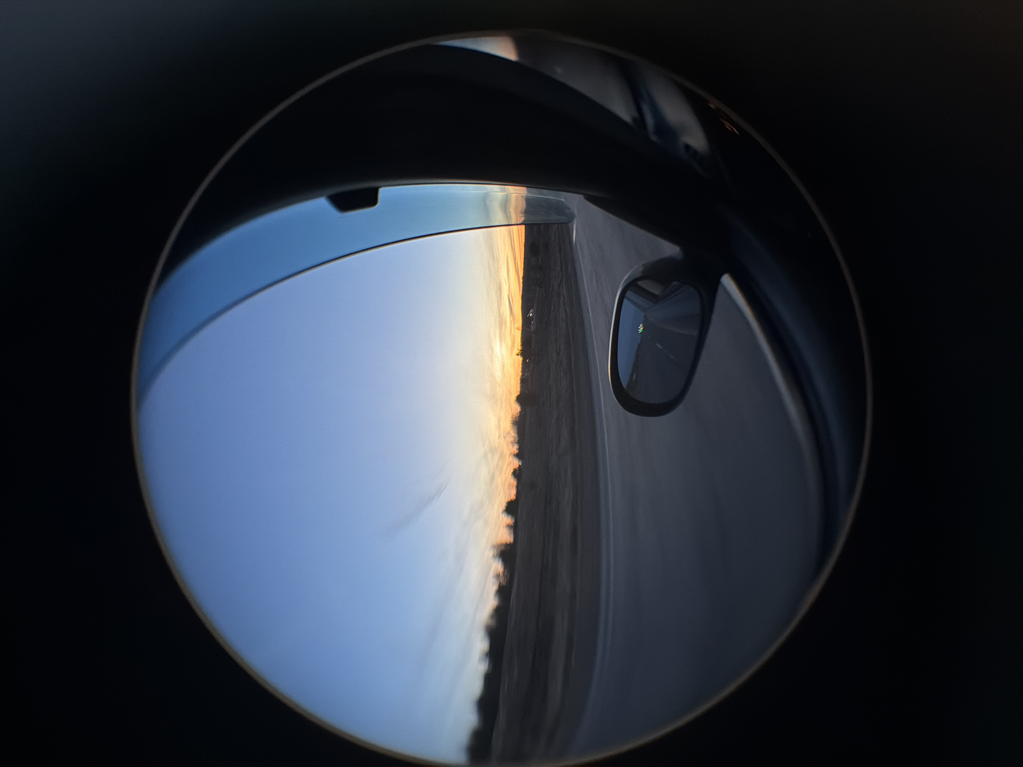 The image shows a distorted view of a sunset landscape, captured through a curved surface that could be a lens or a specialized mirror. The lower half of the image is dominated by a blue sky transitioning into a horizon line marked by golden light, suggesting the sun is setting or rising. The ground is discernible as a flat, grey surface, possibly a road or a field, with darker vertical features, possibly trees or vegetation, separating the sky from the land. The upper part of the image reveals details of a vehicle's interior: the edge of the roof, part of the door frame, and the side-view mirror reflecting the passing landscape. The scene is contained within a circular frame against a black background. The image does not contain any discernible human figures or interactions. The location details of Kuregovo, Russia, are not directly discernible within the image.