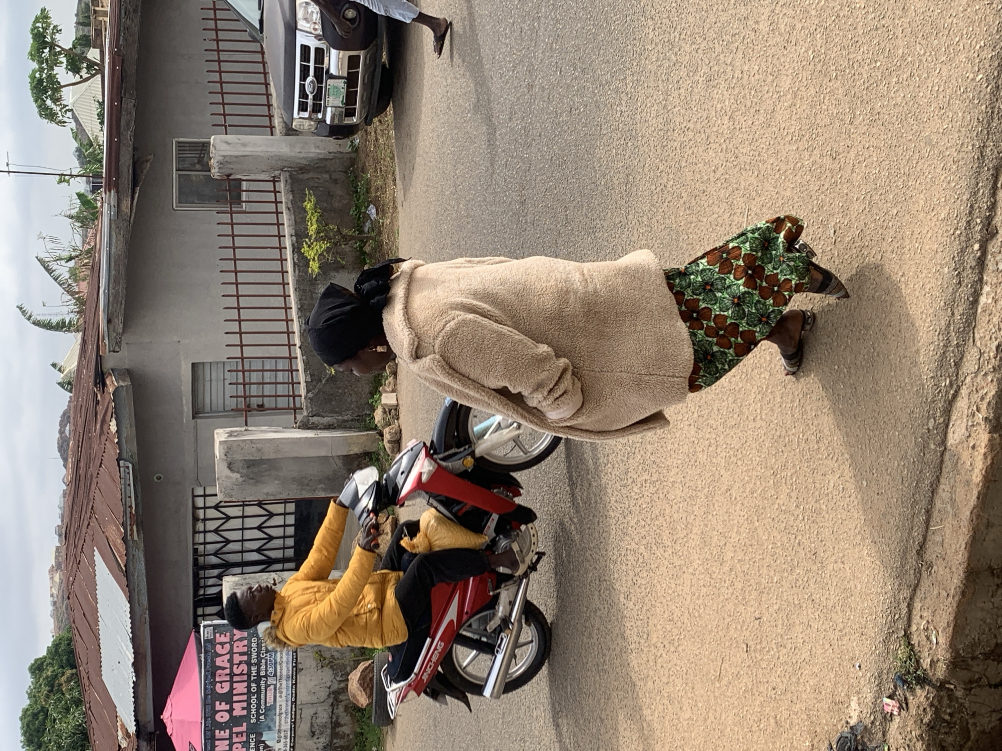The image depicts a daytime street scene in what appears to be an urban or semi-urban area, likely in Jos, Nigeria.
In the midground, a person wearing a yellow jacket and black pants is seated on a red and black motorbike, which has "JINCHENG" written on its side. This individual is positioned on the left side of the street. To their right, a person wearing a light brown, fluffy jacket, a black head covering, and a vibrant green and brown patterned skirt is walking on the asphalt road, passing the motorbike.
In the background, several structures line the left side of the street. These include simple buildings with corrugated metal roofs. A notable feature is a sign affixed to one of these buildings, displaying information about a ministry. The legible text on the sign reads: "NE OF GRACE PEL MINISTRY," "SCHOOL OF THE SWORD. (A Community Bible Class)," and "THUR 4:30PM." Below this, there are contact details and social media handles like "@Othroneofgrace" and an address "F Cerpers' Lodge, Dadin Kowa First."
On the right side of the street, a dark-colored SUV or car is partially visible, parked or moving slowly. The road surface is asphalt, and the lighting suggests a clear, sunny day. The scene captures everyday activity in a local setting, with people and vehicles on the street amidst residential or commercial buildings.
