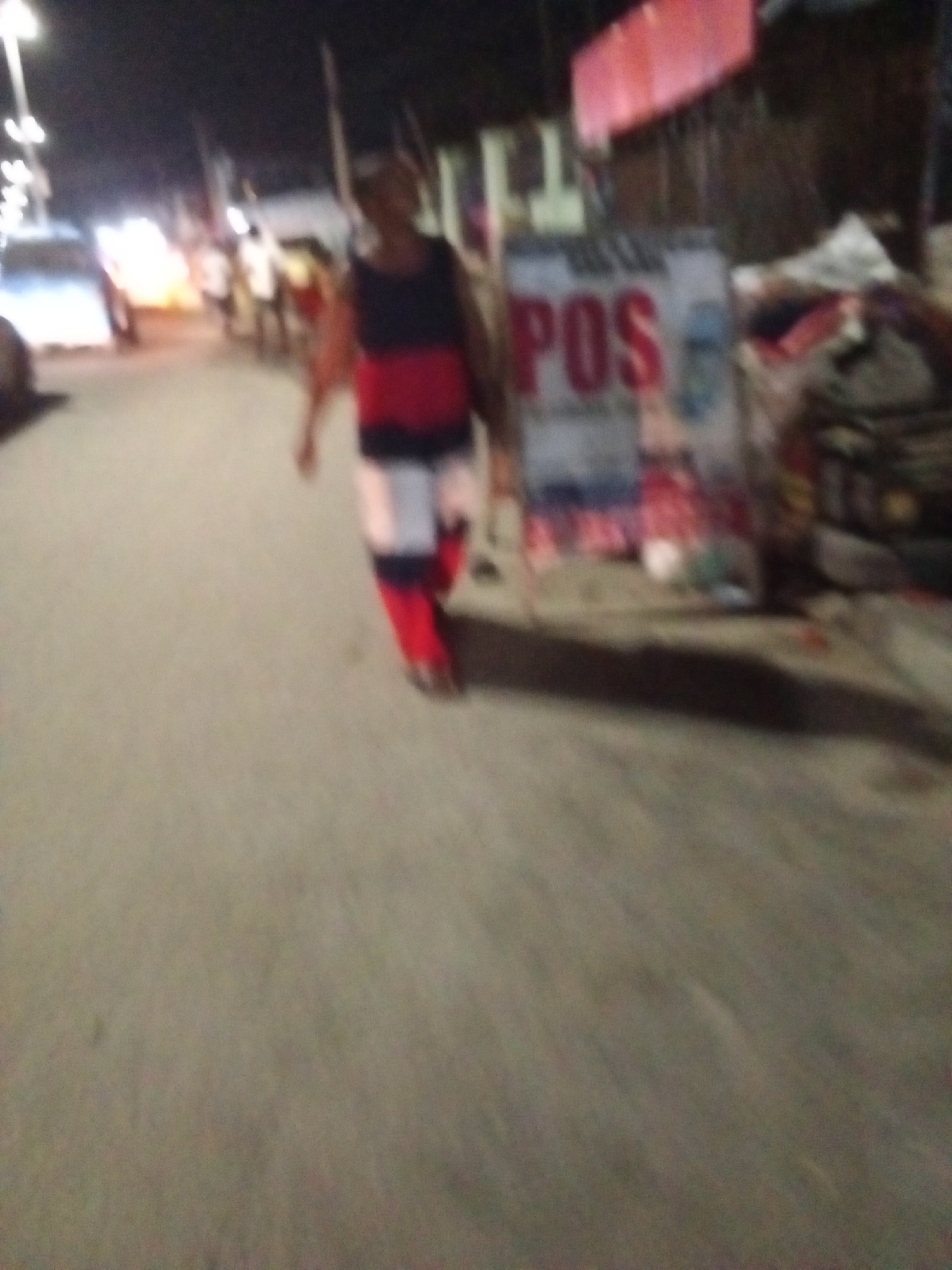 A blurry, low-light photograph appears to be taken in Ado, Nigeria, given the presence of a "POS" (Point of Sale) sign, common in Nigerian commercial settings. A man in a dark tank top, red t-shirt, white shorts, and red leggings walks along a street. Several other figures are visible further down the street, under streetlights. A jumbled pile of objects is on the roadside near the "POS" sign. The general setting is an urban street at night.