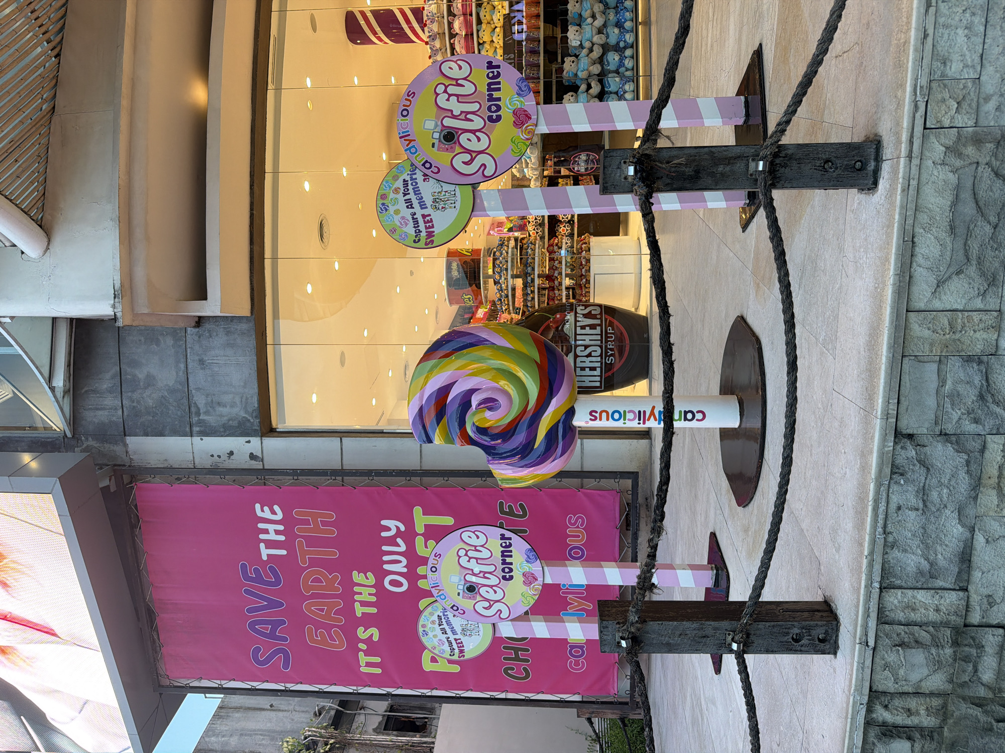 This is an outdoor shot of the storefront of Candylicious, a retail store. The store appears to be located in a tourist-oriented area, likely a shopping mall. The signage visible promotes a "Selfie Corner" and the slogan "Save The Earth." The image captures colorful and whimsical decorations, including lollipop sculptures, candy-themed signs, and large Hershey's Syrup bottle. The facade is predominantly glass, allowing visibility into the interior, which is filled with candy-themed merchandise and displays. There is rope in the image.
Based on the presence of Candylicious and its tourist-oriented signage, it's highly probable that this image was taken in Kuta, Indonesia.