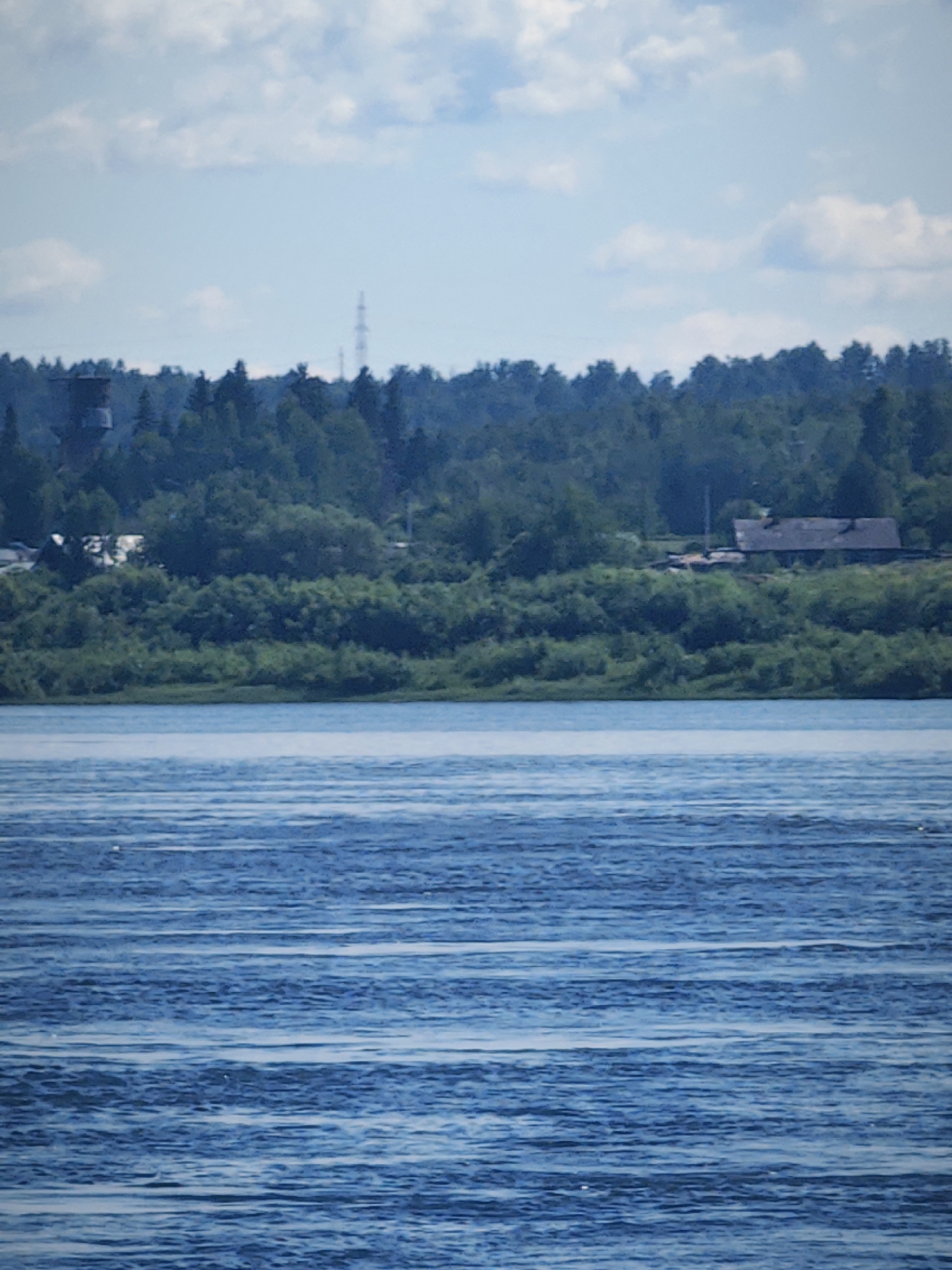 The image shows a wide river with a heavily wooded bank. The river is calm with gentle ripples. The bank is lined with dense green trees and some bushes, creating a lush, natural landscape. In the background, there are a few structures visible through the trees, including a tall tower and a building with a dark roof. The sky is partly cloudy with patches of blue sky visible. The overall scene is tranquil and natural, suggesting a rural or semi-rural setting. It is possible this location is in or near the city of Абалаково, Russia, as indicated.