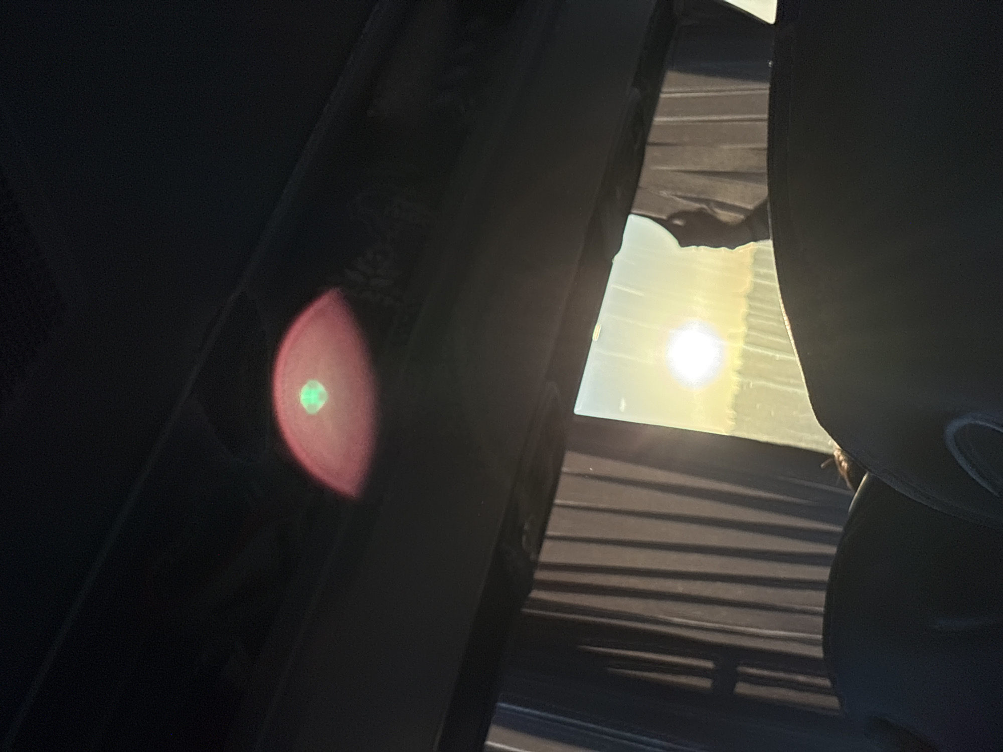 The image shows a view from inside a vehicle, likely a car. The sun is visible through a window, casting strong shadows. The interior of the car is dark, with parts of the seats visible. A circular reflection with a green spot is visible on the car's interior. The exact location in Khvalynsk, Russia, is not discernible from the image.