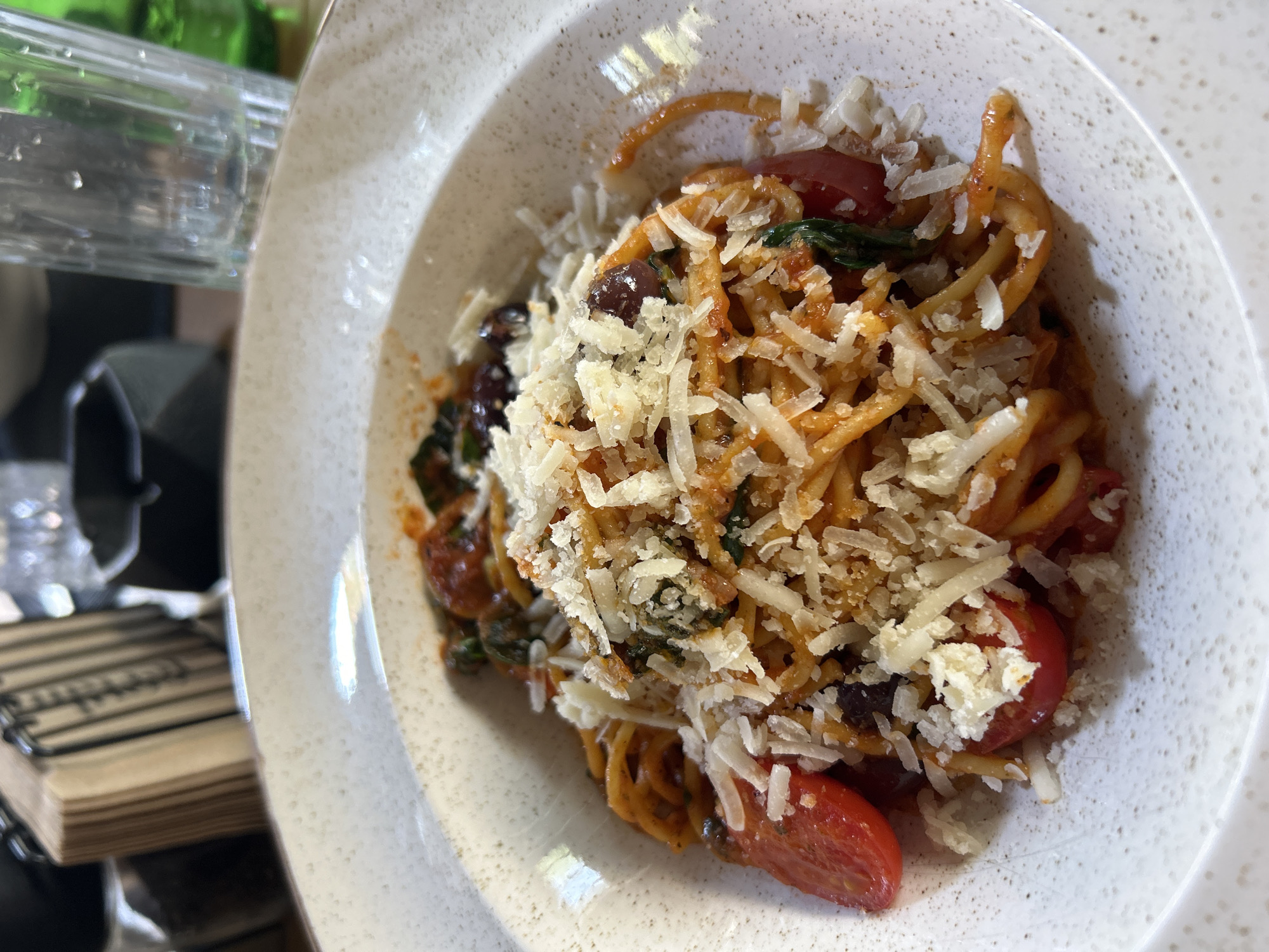 A close-up shot shows a bowl of spaghetti with a red sauce, garnished with shredded cheese, halved cherry tomatoes, spinach, and olives. The bowl is off-white with speckles. A clear glass container and a wooden object are partially visible in the background. The location is Sofia, Bulgaria.
