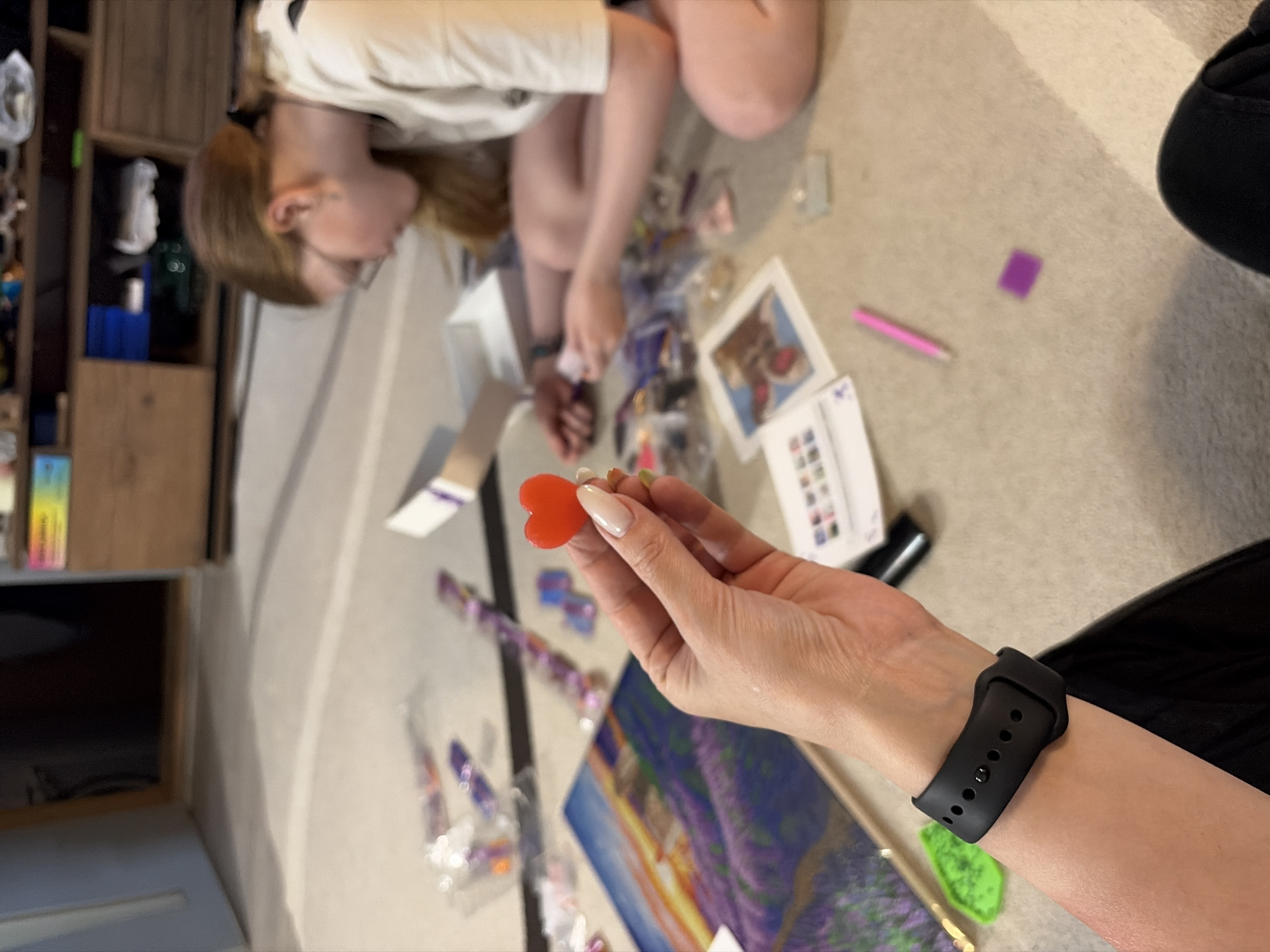 A partially blurred overhead shot shows two people, one holding a heart-shaped object, working on a craft project on a light-colored carpeted floor. A fair-skinned female with blonde hair sits in the upper left of the frame, wearing a white top and glasses, and appears to be working with small plastic bags and a white box. To her right, a hand, likely belonging to the photographer, holds a small red heart-shaped object. The hand also sports a black smartwatch on the left wrist. Spread out around them are various crafting supplies, including a painted canvas of a lavender field, small colored plastic pieces, and a chart. A wooden shelf is visible in the far left. The location is unspecified, and there is no information about the location in the city of Chaikovsky, Russia.