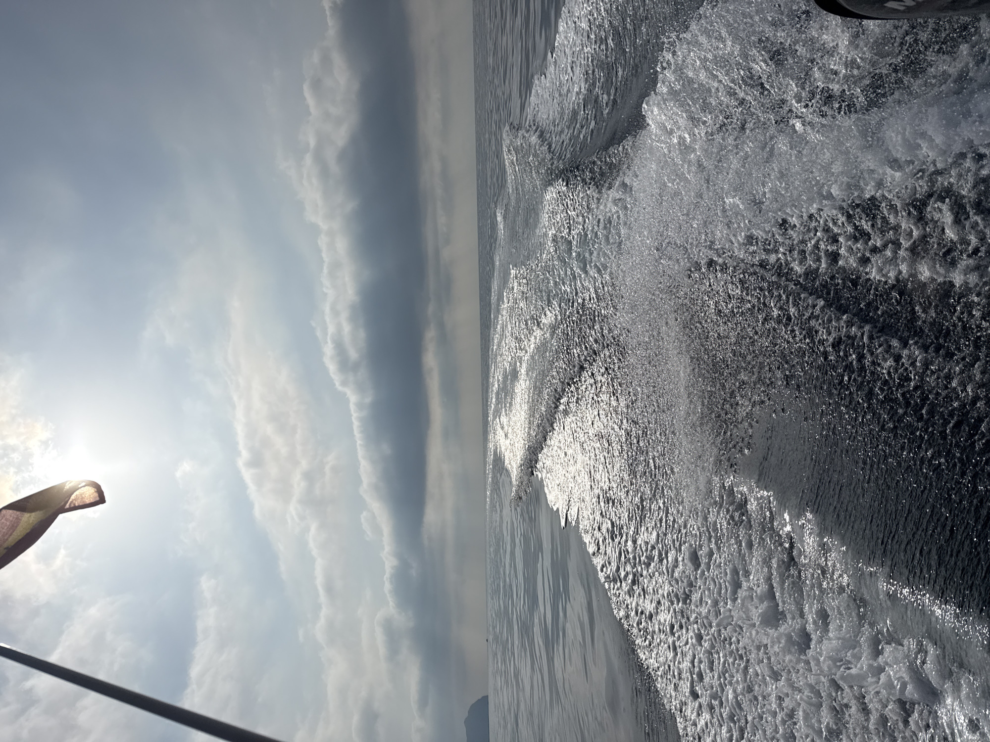 A rear view from a boat shows a wake trailing behind on a calm sea under a partly cloudy sky. A flag, possibly the Spanish flag due to the colors red and yellow, is visible at the top left, attached to a flagpole. Sunlight streams through a gap in the clouds, creating a bright halo effect. In the distance, a landmass, likely an island or coastline, is faintly visible on the left horizon. The overall scene suggests a journey at sea, possibly near the coast of Águilas, Spain, given the potential flag and maritime setting.