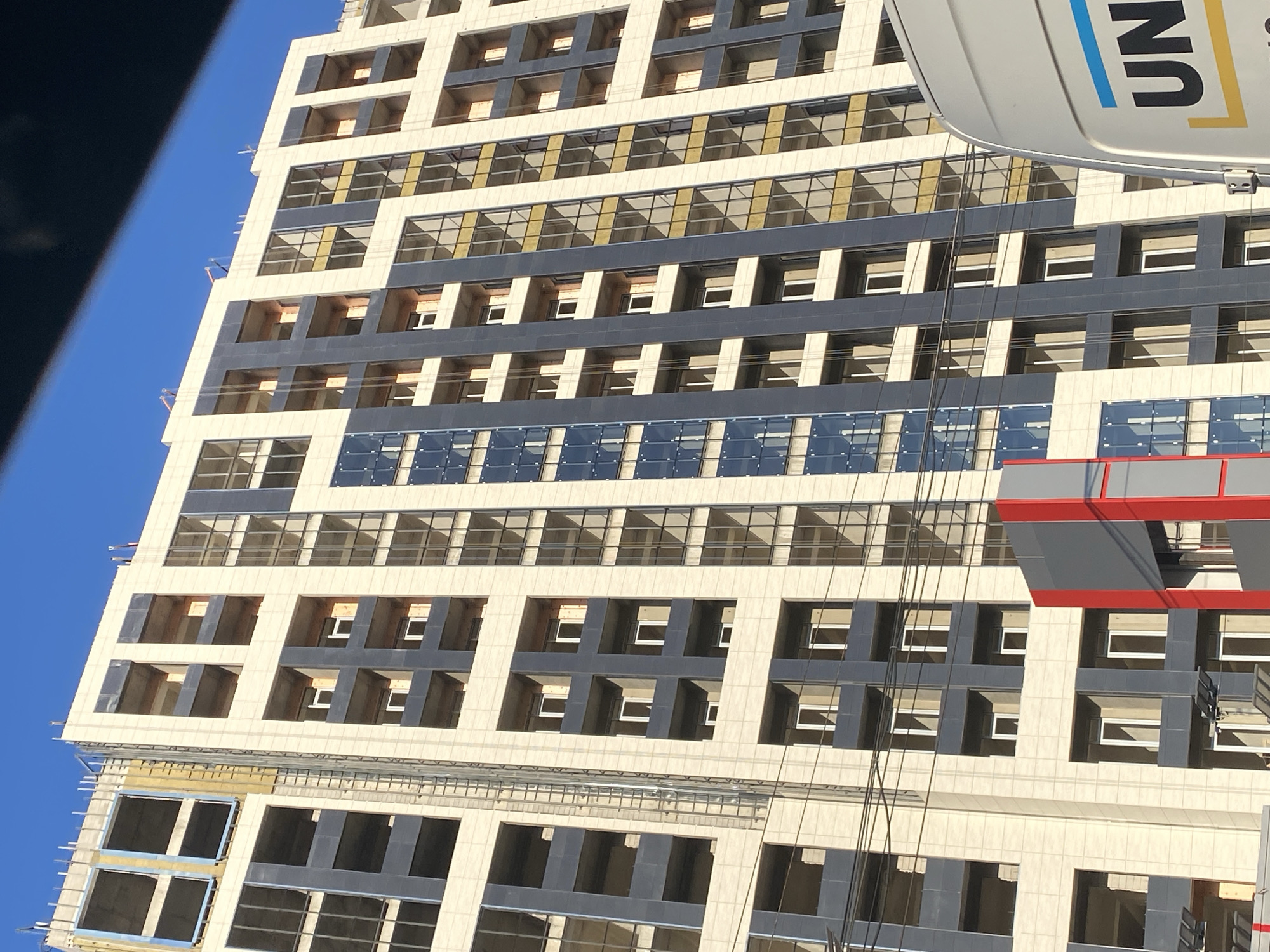 This media file is an image of a modern skyscraper under construction in Yerevan, Armenia. The building features a repeating pattern of light-colored facade panels interspersed with dark grey rectangular window openings. Some of these openings are complete with glass, while others appear to be unfinished, showing the internal structure.
The skyscraper is tall and imposing, with a clear blue sky as the backdrop, suggesting it is daytime with fair weather. The perspective is from below, looking upwards, which emphasizes the height and scale of the building.
A notable object in the upper right portion of the image is part of a white structure, possibly a crane or a piece of equipment, with the letters "UN" in black and blue, and yellow graphic elements, visible on it. This suggests the construction may be related to or overseen by an organization with the "UN" designation.
The building's design is contemporary, with clean lines and a geometric aesthetic. The construction appears to be in an advanced stage, with many floors already clad, but some areas still showing exposed concrete and structural elements. The overall impression is one of urban development and progress in Yerevan. There are no people visible in the image.