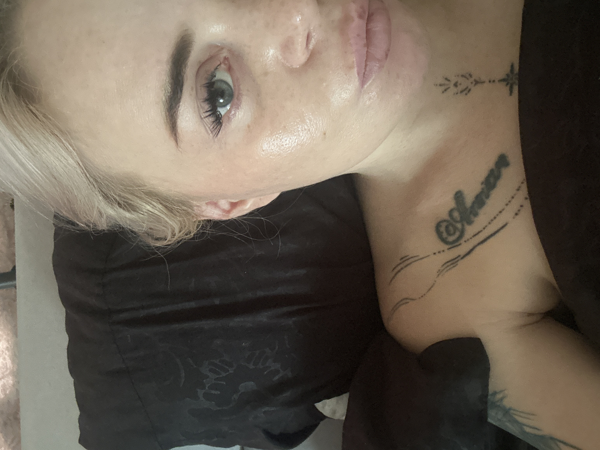 An upper body shot of a fair-skinned woman with blonde hair is captured in what appears to be a bedroom. She is lying down, with her head resting on a dark pillow. Her face is turned towards the camera, with her left eye and part of her nose and lips visible. Her skin has a dewy appearance, with subtle blush on her cheeks and what looks like light eye makeup. Her lips are a soft pink. She is wearing a dark, possibly black, top with a textured pattern. A tattoo is visible on her chest, partially obscured by her top. Another tattoo is visible on her left arm, near her shoulder. The background is slightly blurred, suggesting an indoor setting. The lighting appears to be natural, possibly from a window.