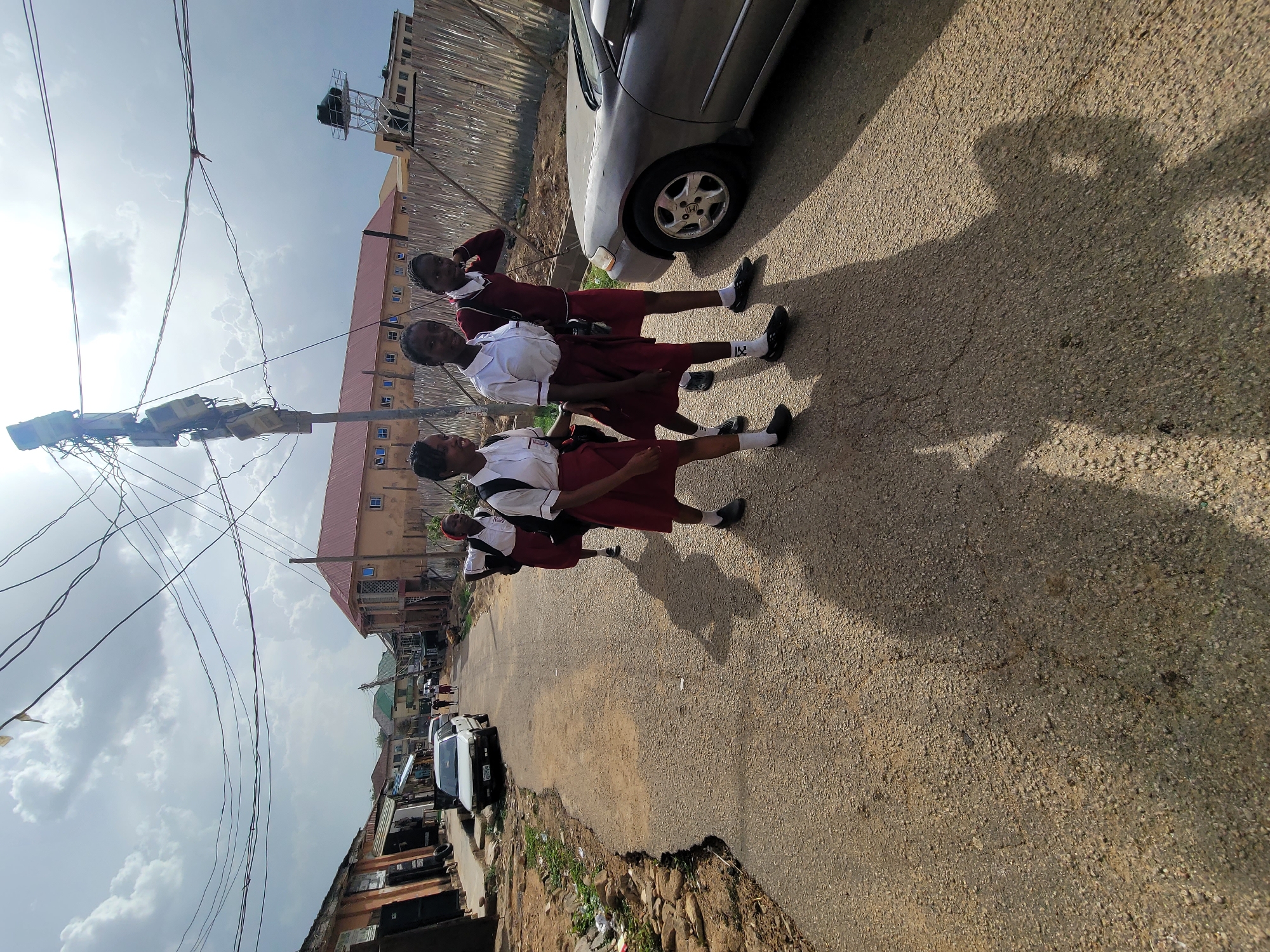 The image depicts a street in Jos, Nigeria. Four schoolgirls in red and white uniforms are walking on the side of the road, next to a parked gray car. An overhead power pole is visible on the left side of the frame, with wires extending across the scene. Buildings and parked vehicles line the street. The road surface is cracked and shows signs of wear. The sky is overcast.