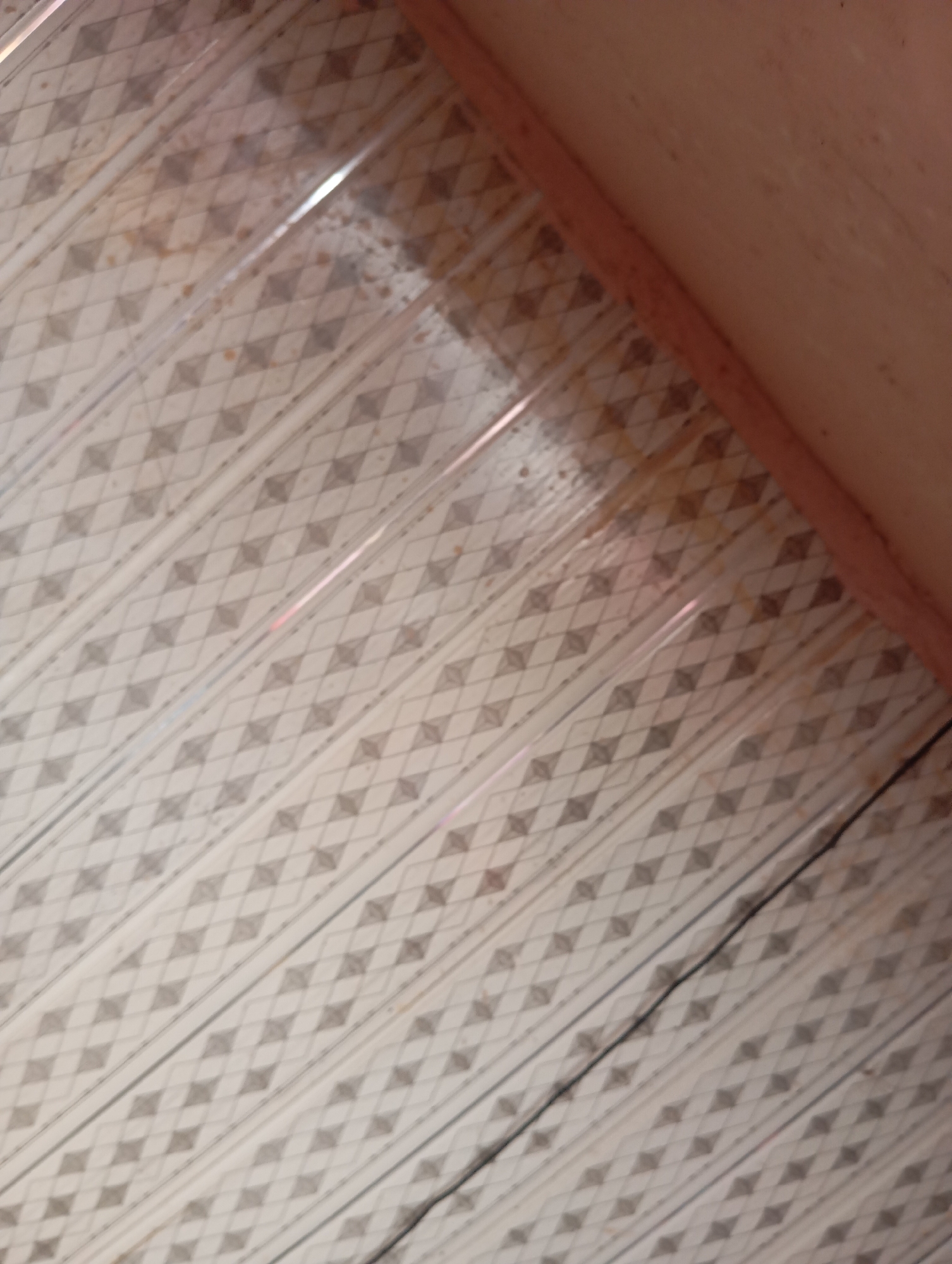 The image shows a close-up, tilted view of a ceiling. The ceiling is made of corrugated plastic sheets with a diamond pattern. There are visible stains and marks on the plastic, suggesting dirt or water damage. A light pink, textured surface, possibly another part of the ceiling or a wall, is visible in the upper right corner. A thin black wire runs diagonally across the patterned surface. The lighting suggests an indoor environment. The image lacks any identifiable landmarks to determine its specific location within Jalingo, Nigeria.