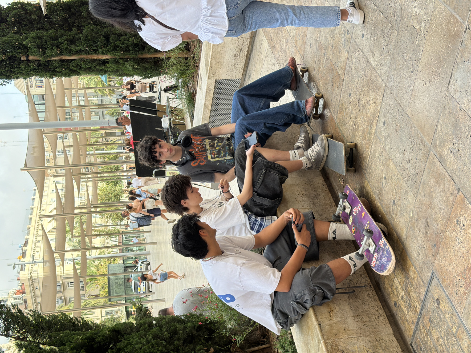 The image captures a street scene in an urban environment, possibly a public plaza. In the foreground, three young men are sitting on a low stone wall, each with a skateboard nearby. The man on the left wears a white t-shirt and grey cargo shorts, with a skateboard featuring a purple deck and stickers positioned in front of him. The middle individual is in a white t-shirt and plaid shorts, holding a mobile phone. The third young man is seated on a skateboard, wearing a graphic t-shirt and dark blue jeans, with headphones around his neck.
Behind them, other people are visible in the background, including individuals walking and some seated. The architecture suggests a European city with buildings featuring light-colored facades and awnings. The ground is paved with large stone tiles. The overall atmosphere is casual and relaxed, suggesting a daytime gathering in a public space.