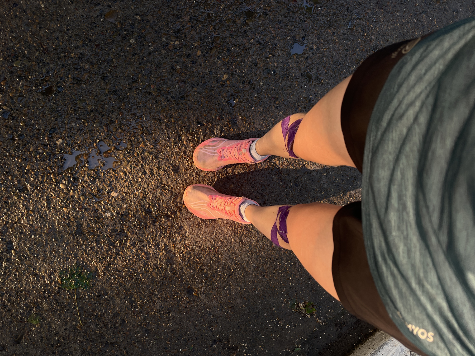 This media file shows a first-person perspective of someone standing on a wet, asphalt surface. The individual is wearing pink running shoes with purple athletic tape around their calves and dark shorts, covered partially by a teal athletic top. The ground appears to be a dark asphalt mixed with small pebbles and has several small puddles reflecting the light. A single green leaf is visible on the ground to the left of the frame. The lighting suggests it is either early morning or late afternoon, with a warm glow illuminating the scene, casting long shadows. The wet ground indicates recent rain or dew. There is text visible on the teal athletic top, reading "AYOS" in white lettering. The overall impression is that of someone preparing for or having just completed an outdoor physical activity, possibly running, in Chaikovsky, Russia.