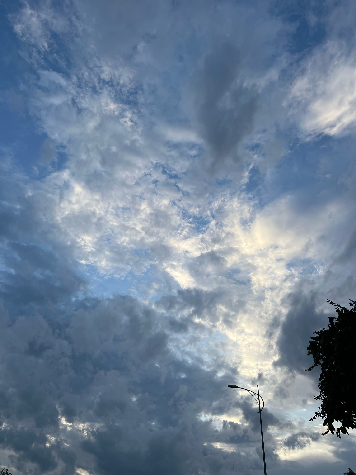 This is a photograph of the sky above Nha Trang, Vietnam. The sky is filled with a mix of dark gray storm clouds and lighter, wispy clouds, with patches of blue sky visible between them. A street lamp with a curved arm is visible in the lower right quadrant of the image, silhouetted against the sky. To the far right, the dark silhouette of tree branches and leaves is visible. The lighting suggests it may be late afternoon or early evening, with the sun partially obscured by the clouds.