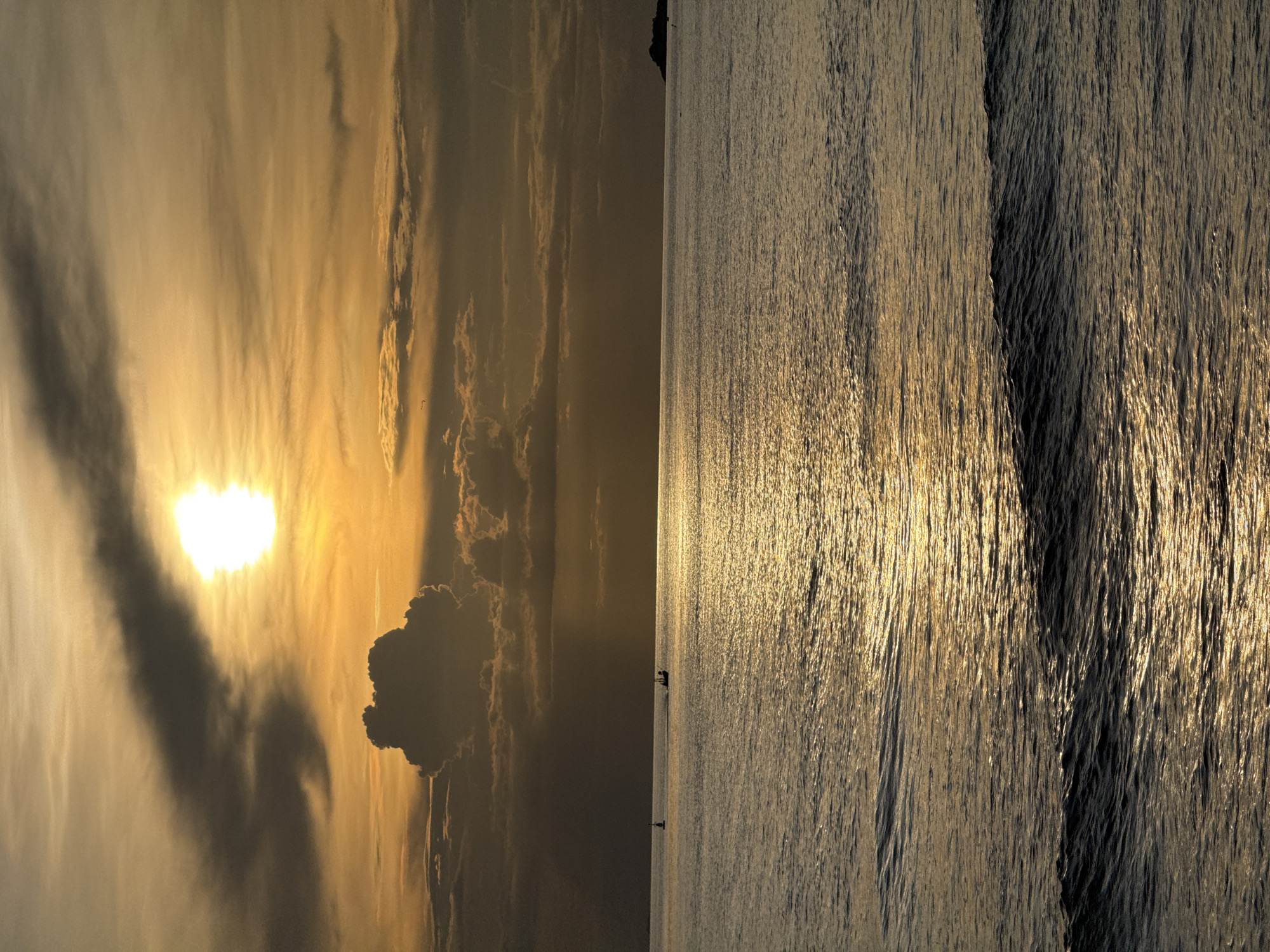 A sunset over the sea is depicted in this image, likely taken from the shore of Koh Phangan, Thailand. The sun, a bright white orb partially obscured by clouds, casts a warm golden glow across the sky and water. The sky is filled with layers of clouds, some dark and dramatic, others wispy and thin. The sea is calm, with gentle waves reflecting the sunlight. In the distance, a small boat is visible, its silhouette dark against the bright water. There are no people visible in the scene. The overall impression is one of tranquility and natural beauty.