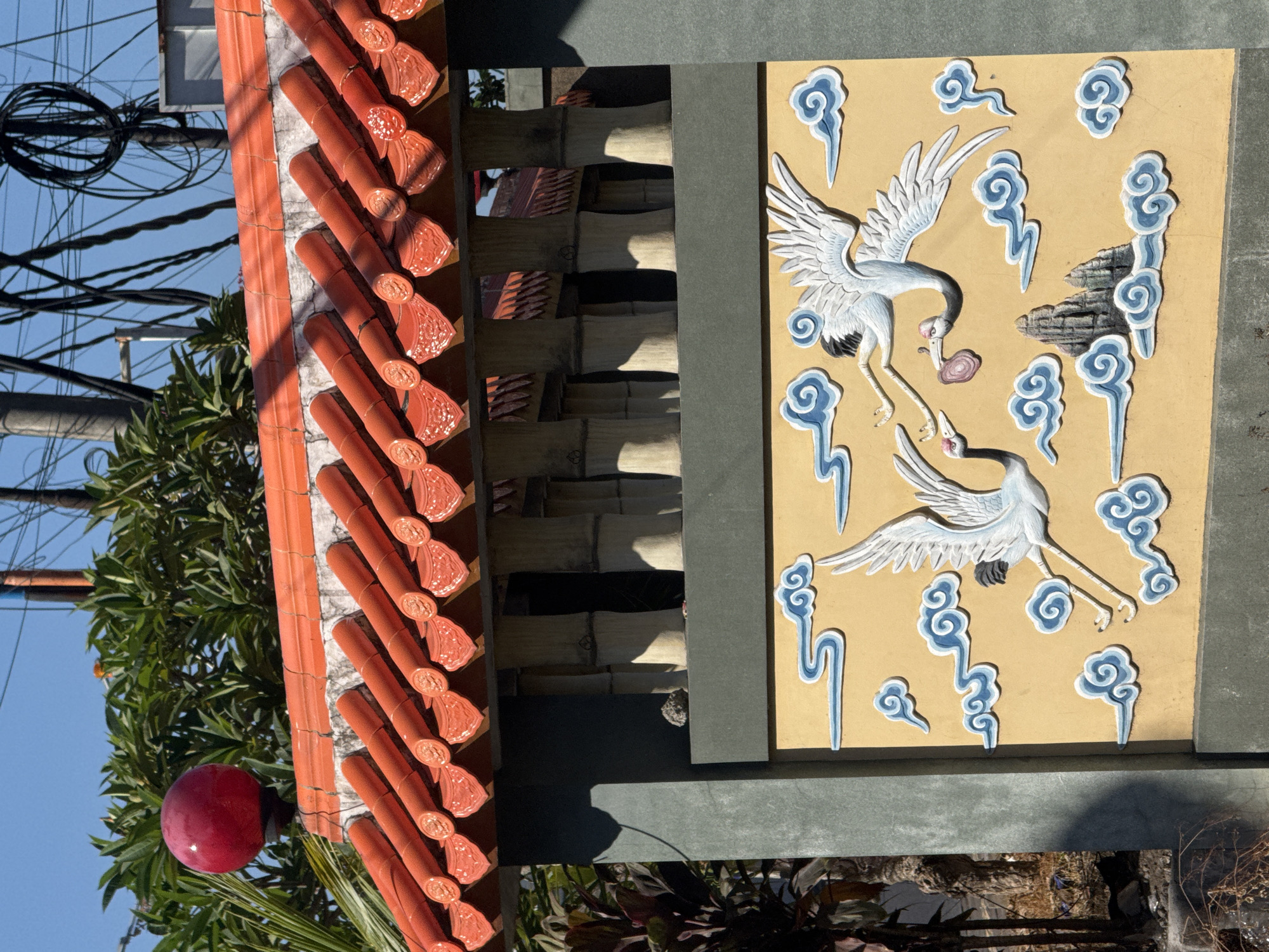 The image shows a section of a traditional Balinese structure, likely a temple or a home compound, located in Denpasar, Indonesia.
The structure consists of a terracotta tiled roof, bamboo paneling, and a painted panel featuring two white cranes amid stylized blue clouds. The terracotta tiles are barrel-shaped and arranged in rows. The bamboo paneling consists of thin, horizontally aligned bamboo stalks. The painted panel is set against a light yellow background with the cranes depicted in a low-relief style.
Additional details include overhead power lines and a red ornamental sphere visible amongst foliage. The scene appears to be photographed in daylight under sunny conditions.