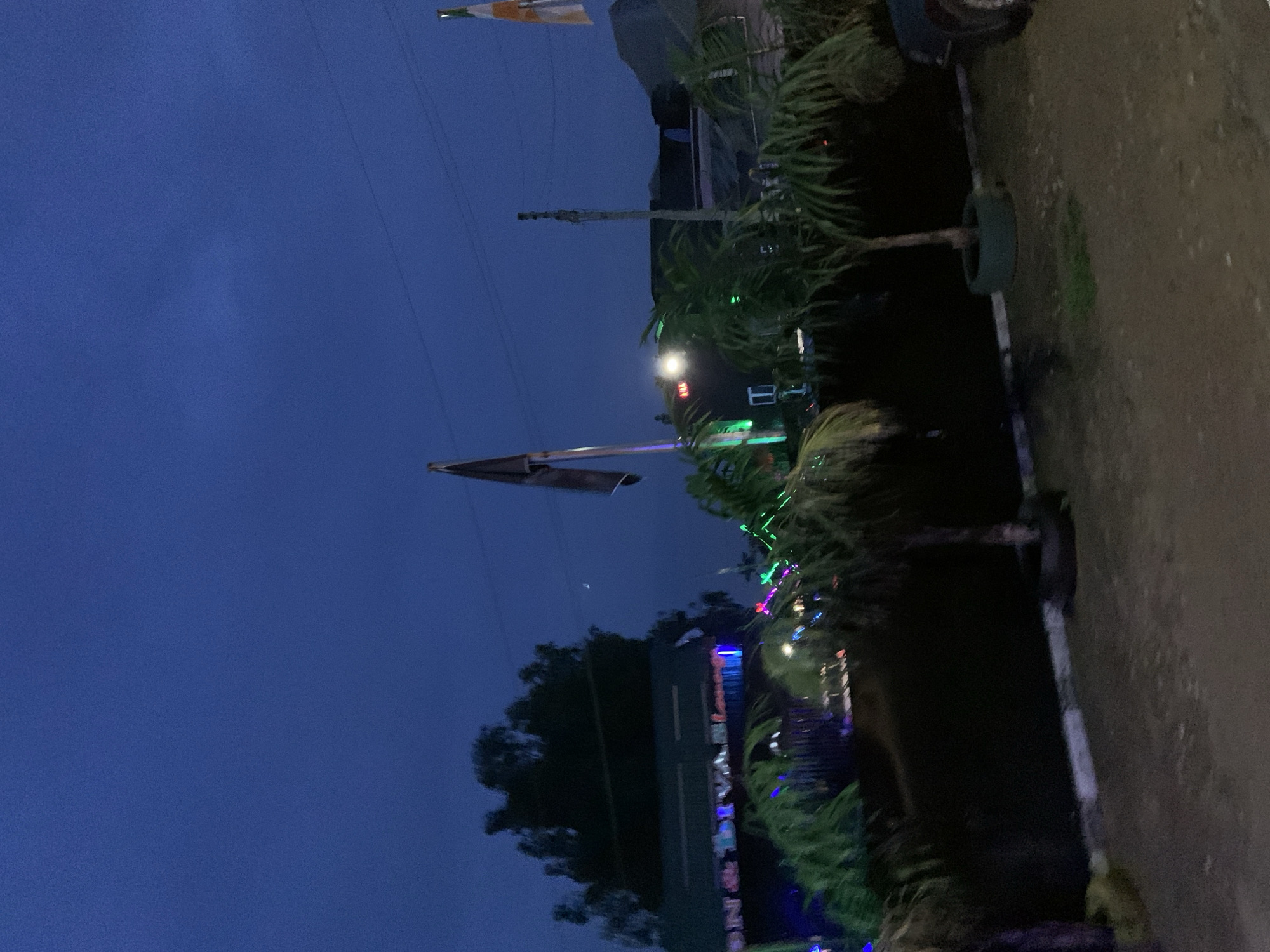 The image depicts an outdoor scene at dusk in Gura Top, Nigeria. In the foreground, a gravelly surface transitions to a darker, possibly paved area with some white curbing. Several potted palm trees are visible, with one on the right planted in a repurposed tire.
In the middle ground, a building with a green roof is illuminated by colorful string lights, some appearing blue and green. The building has signs with lettering that is partially obscured but seems to include "ON & DIVINE". There are also vertical structures resembling poles, one with a dark flag.
Further back, another building, darker in color, has a few visible lights, including a red one. The sky above is a deep blue, characteristic of twilight, with a faint crescent moon visible. There are overhead wires stretched across the scene. The overall impression is that of an outdoor establishment, possibly a restaurant or entertainment venue, decorated for the evening.