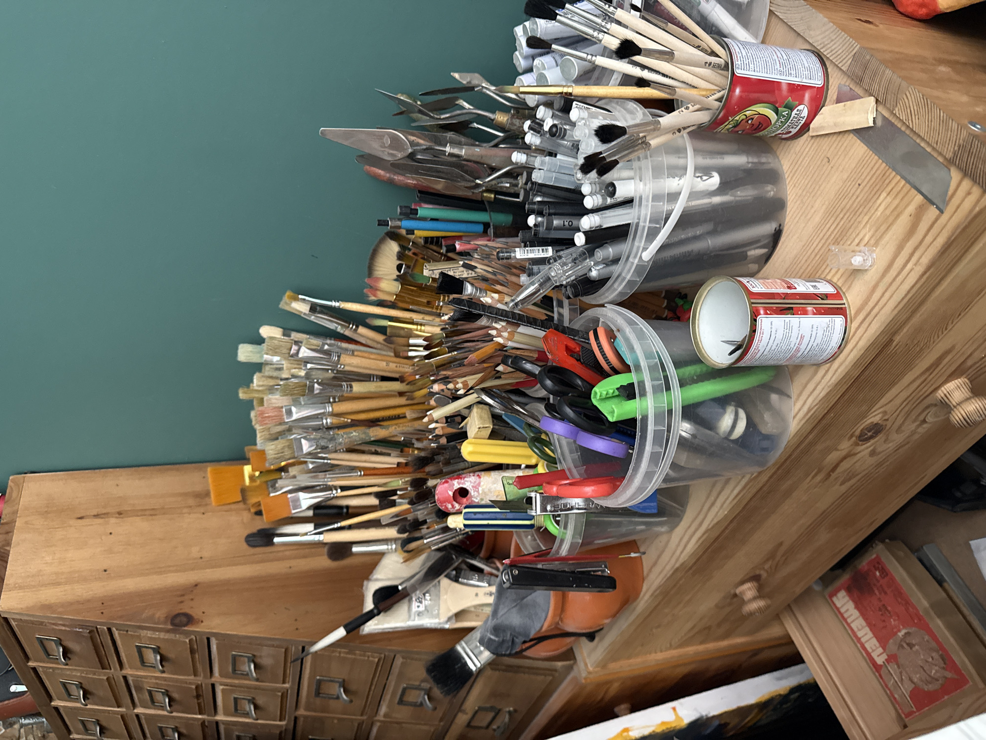A cluttered wooden shelf in an art studio displays a variety of art supplies. In the background, a tall wooden cabinet with multiple small drawers is visible. The foreground is dominated by numerous containers filled with paintbrushes, pencils, pens, and art tools such as palette knives and tweezers. Some of the containers are clear plastic buckets, while others are repurposed tin cans, one of which features a label with Russian text and an image of tomatoes. A partially visible painting on canvas is leaning against the left side of the shelf. The overall impression is one of a working artist's space, likely located in a private residence or studio, though the specific location in Votkinsk, Russia, cannot be determined from the visual information provided. There are no people or animals present in the scene.