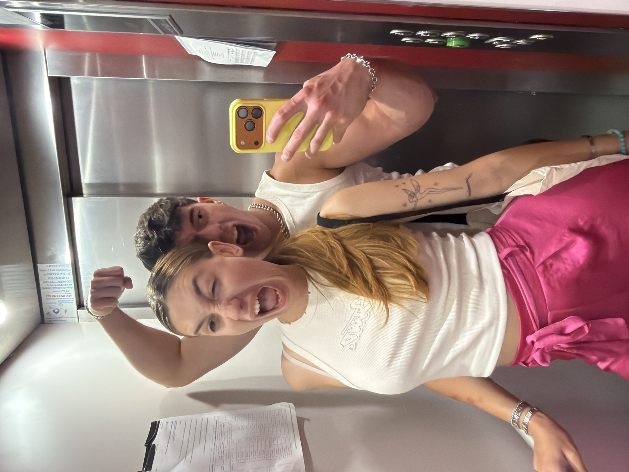 In an elevator, a young woman with blonde hair pulled into a ponytail and a white tank top is taking a selfie. She is winking and has her mouth open. She is wearing a pink satin skirt, and several bracelets on her left wrist. Behind her, a young man with dark curly hair is also in the frame, flexing his bicep for the camera, wearing a white tank top and a silver chain necklace. The elevator doors are silver and red. A yellow phone is held by the man as he takes the selfie. The image does not provide specific details about the city of Sofia, Bulgaria.