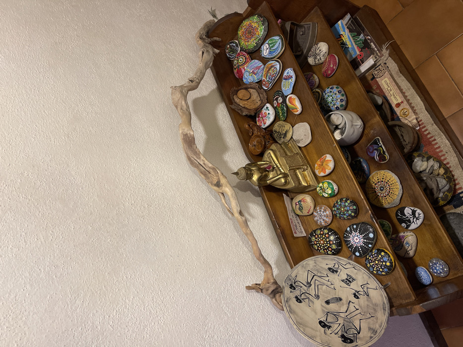 The image presents an indoor decorative display, primarily focused on a wooden multi-tiered shelf unit mounted against a light-colored, textured wall. A long, intricately twisted piece of natural driftwood extends vertically along the wall, adding an organic and rustic element to the scene.
The wooden shelf is densely populated with a diverse collection of items. The most prominent subjects are numerous painted stones, showcasing a wide array of designs. These include intricate mandala patterns in various colors, vibrant floral motifs, abstract shapes, depictions of owls, and symbolic imagery such as Hamsa hands. A golden Buddha statue, appearing to be in a reclining or meditative pose, is positioned centrally on one of the lower shelves. Also visible are a round, light-colored ceramic plate adorned with dark, stylized figures that seem to be dancing or engaged in a ritual, a white spiral-shaped ceramic sculpture, and a small wooden bowl. Further down, more painted stones feature designs like a coiled snake and abstract patterns.
The scene suggests a curated personal collection within a home setting, reflecting interests in art, spirituality, and nature. The ambient lighting is typical of an indoor environment. No specific activity or event is taking place; it is a still life composition. On the upper shelves, a blue and white package has the text "TEX" clearly visible. Below it, a white rectangular packet displays "CORAL BLANCO" along with a small red graphic. The overall aesthetic is eclectic and peaceful, located in Dosso (Ciappanico), Italy.