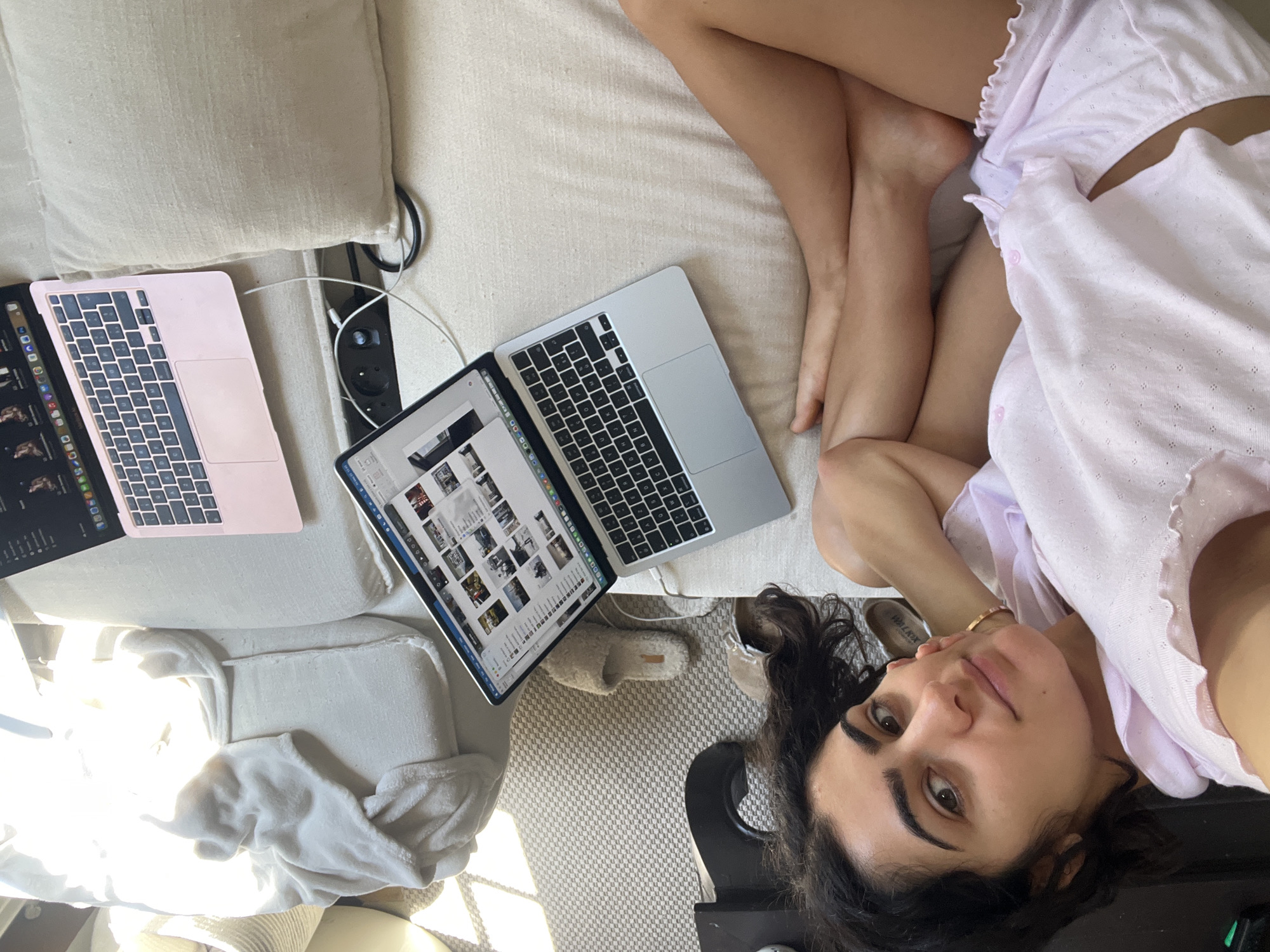 A woman in pink pajamas lies on a light beige couch or bed with several electronic devices. Two laptops and a tablet are visible on the left side of the image. The woman's legs are crossed and bent towards her, while her head is positioned in the lower right corner. A black object is visible beneath her head, possibly part of the furniture. The scene appears to be indoors. There is no indication of the city of Paris.