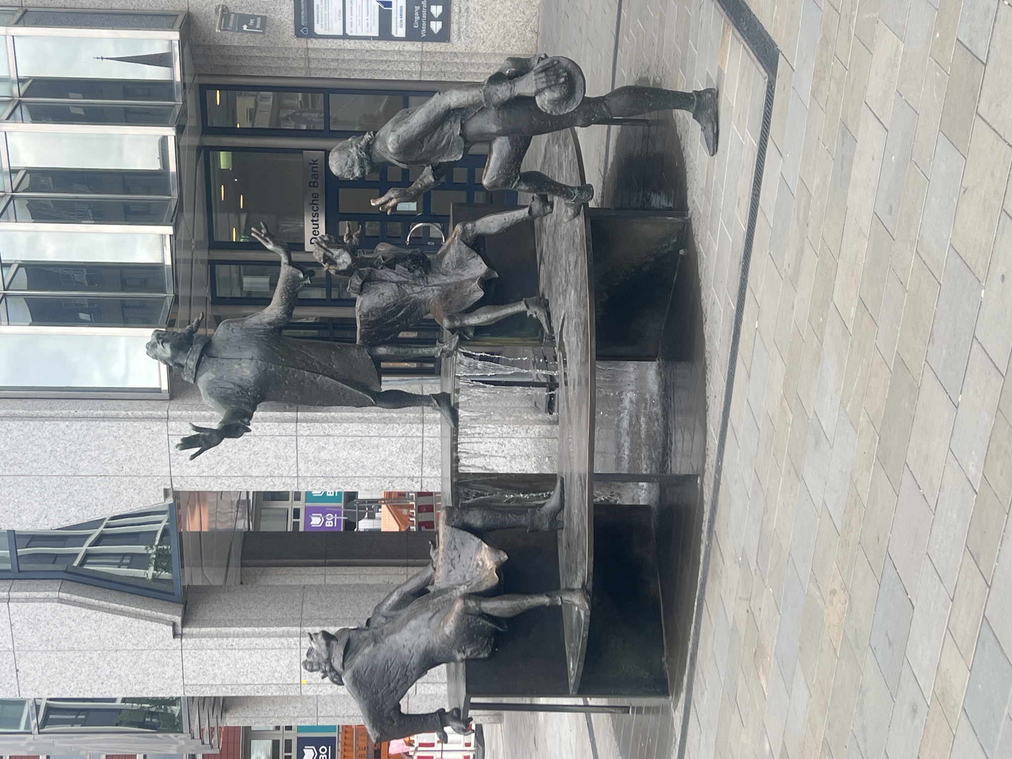 This image shows a bronze sculpture in the city of Bochum, Germany. The sculpture depicts four figures interacting around a fountain. One figure, dressed in a long coat and hat, stands with their arms raised and hands open, as if addressing the others. To their left, a figure is seated on a ledge, looking upwards with their mouth open. Another figure, also seated, is partially obscured by the fountain's water. The fourth figure, standing and holding a hat, gestures towards the seated figure. The figures are all rendered in a realistic style. Behind the sculpture, the facade of a building with large glass windows is visible, with a sign that reads "Deutsche Bank." The ground is paved with large, rectangular stone tiles.