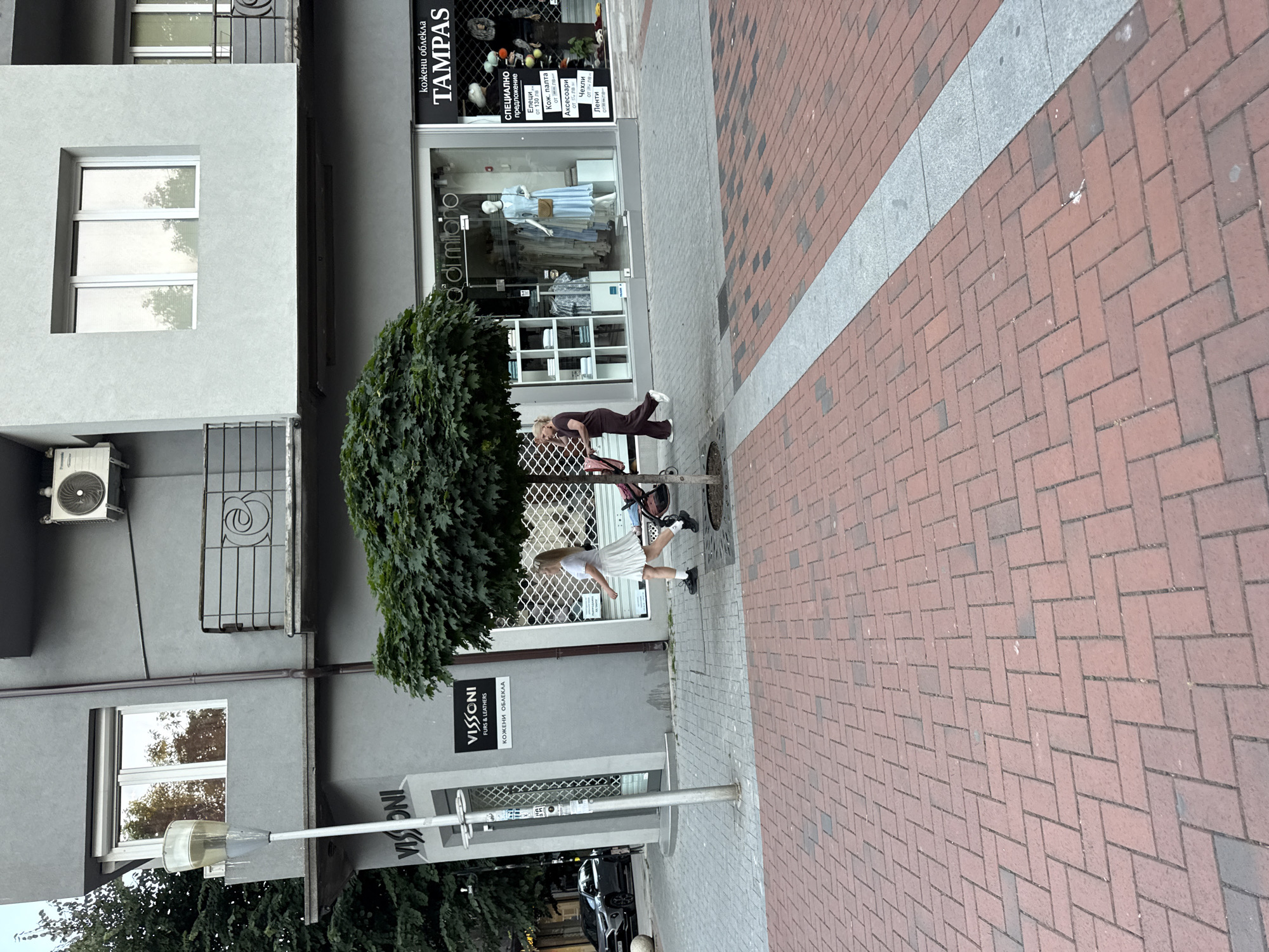 The image shows a street scene in front of shops. Two women are visible, one in the foreground wearing a white skirt and top, and another in the background wearing a dark outfit. They appear to be walking or posing on the sidewalk, which is paved with red brick. A large, leafy tree is also a prominent element in the scene. Behind the tree, there are shop windows displaying clothing and mannequins. Signs with text in what appears to be Cyrillic script are visible on the shop fronts, including "TAMPAS" and "VISSONI FURS & LEATHERS". The time of day appears to be daytime, and the weather seems to be overcast or at least not brightly sunny, judging by the lighting. The overall impression is of a typical urban street with commercial activity.