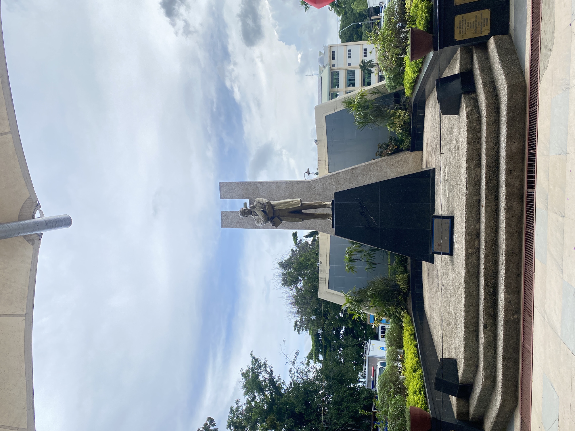 The image shows the Rizal Shrine in Davao City, Philippines. A bronze statue of Jose Rizal stands on a tiered concrete and granite monument. Behind the statue, a tall, angular structure with a dark granite base rises into the cloudy sky. The monument is surrounded by lush greenery, including manicured bushes and potted plants. In the background, a modern building with multiple floors is visible, as are trees and a few vehicles. The area appears to be a public space, possibly a park or plaza, designed to honor the national hero.