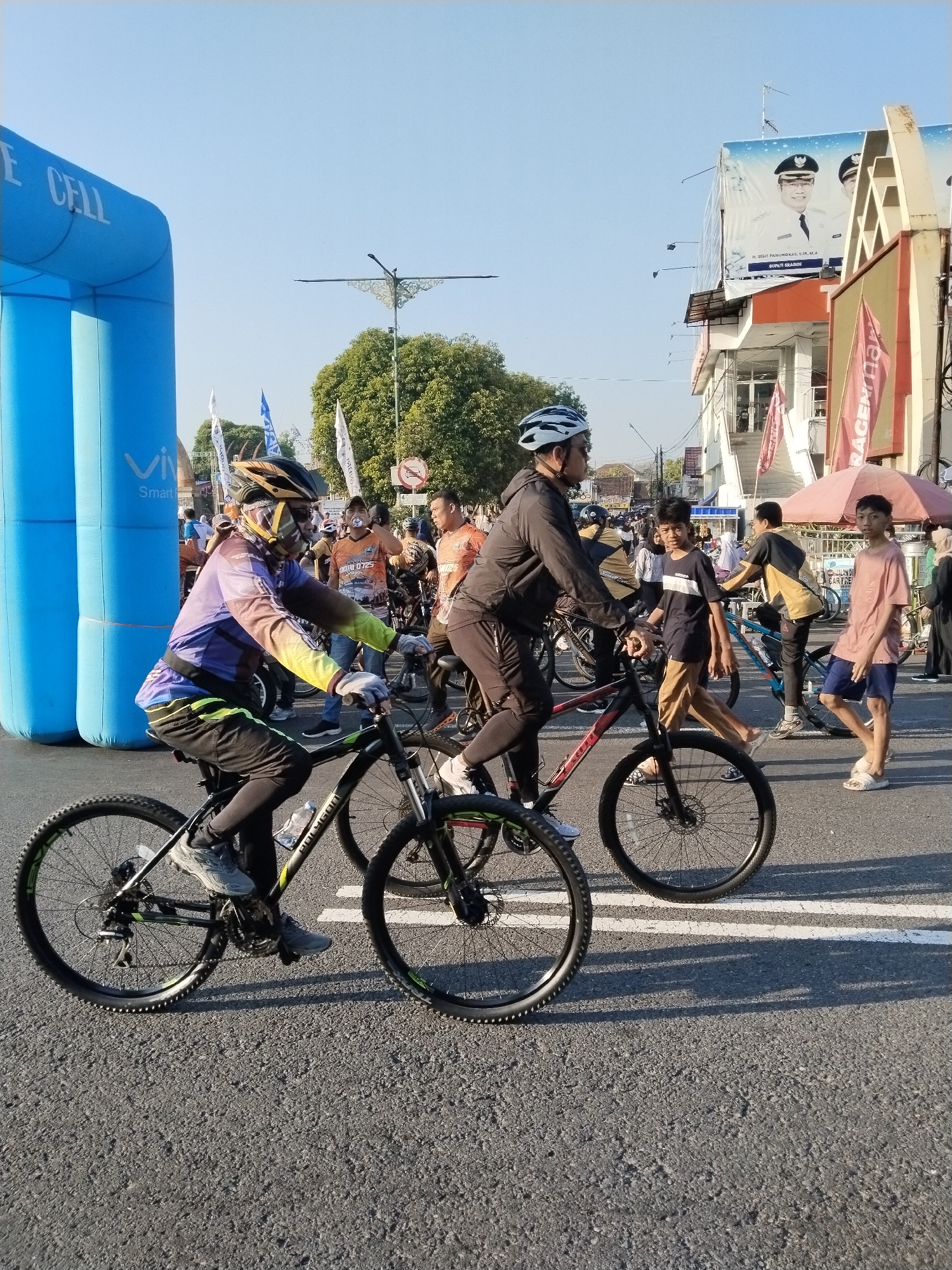 The image depicts an outdoor daytime scene on a paved asphalt road in the city of Sragen, Indonesia. Two individuals are cycling prominently in the foreground, moving from left to right. The cyclist on the left wears a multi-colored long-sleeve cycling jersey (purple, orange, yellow-green gradient), black cycling pants, a black and yellow helmet, a face mask, and gloves, riding a black Polygon mountain bike with green accents. The cyclist on the right wears a dark grey long-sleeve jacket, black cycling pants, a white helmet, and sunglasses, riding a black Polygon mountain bike with red accents.
In the midground and background, a moderate crowd of people is visible, walking, standing, and cycling, suggesting a public event. On the far left, a large blue inflatable arch is partially visible, bearing the text "E CELL" and "Vivo Smart". Buildings with multiple stories line the right side of the street. A large billboard affixed to one of these buildings displays portraits of two men in official attire, with legible text identifying one as "H. SIGIT PAMUNGKAS, S.I.P., M.A., BUPATI SRAGEN," confirming the location as Sragen and the person as its Regent.
Further elements include various white and blue flags, an ornate decorative street light fixture in the upper center, green trees, a red circular traffic sign indicating restricted vehicle access (a car with a diagonal line), and a red market umbrella. The scene is illuminated by bright sunlight, casting visible shadows on the asphalt road, which features white painted lines.