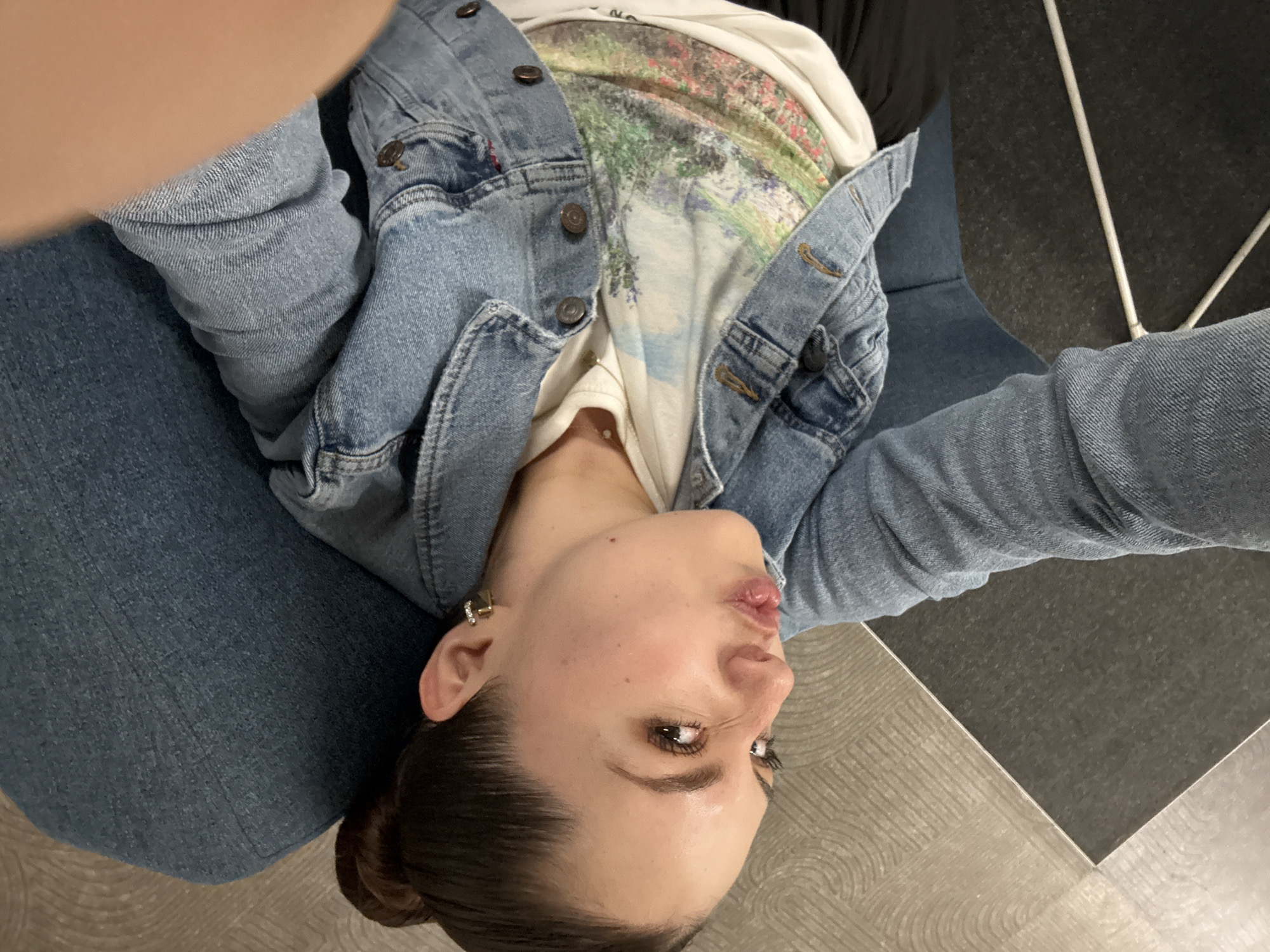 A woman wearing a denim jacket and a t-shirt with a landscape design is shown from a high angle. She is sitting on a blue upholstered chair. The floor is light gray with a dark gray square tile. Part of a white chair leg is visible in the upper right corner. The photo appears to have been taken indoors. The city is not discernible from the image.