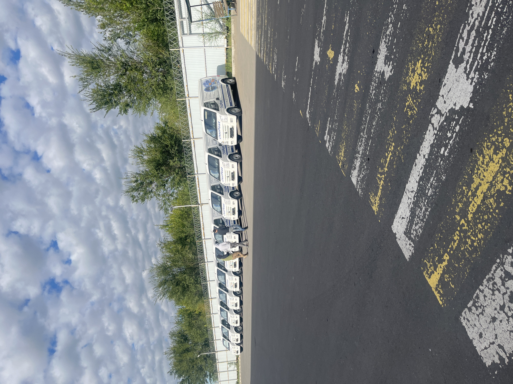 The image depicts a row of white delivery vans parked in a lot, likely a depot, under a cloudy sky with patches of blue. The vans are branded with the logo "OazPost". In the foreground, a pedestrian crossing is marked with faded yellow and white paint on the asphalt. Three individuals are visible walking away from the camera in the midground, between the parked vans and the fence. Behind the vans, there is a white metal fence topped with barbed wire, and beyond that, a line of green trees. The location is identified as the city of Astana, Kazakhstan.