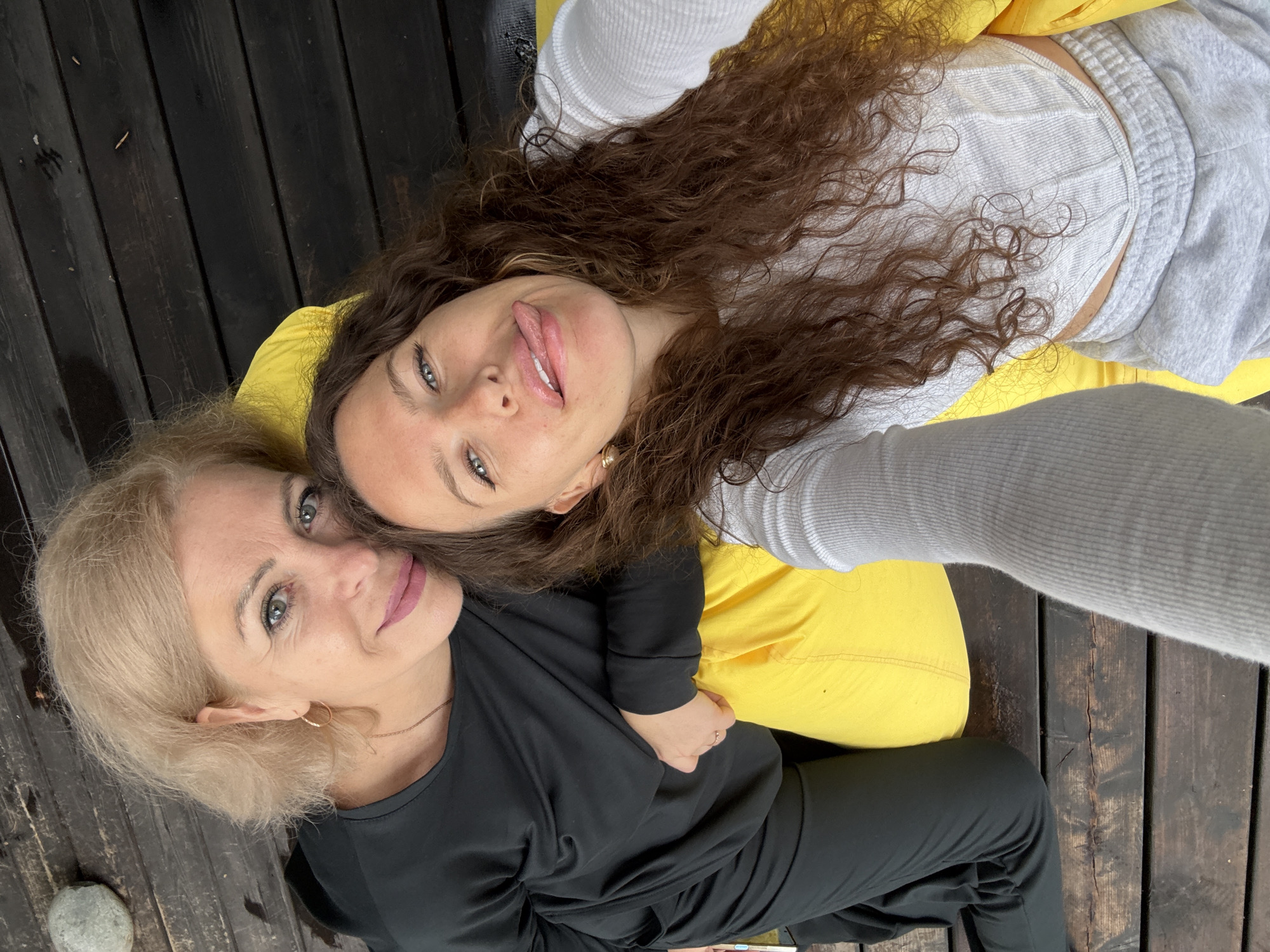 Two women are posing for a selfie on a wooden deck. The woman on the left has blonde hair and is wearing a black outfit. She is looking at the camera with a slight smile. The woman on the right has long, curly brown hair and is wearing a white top and grey sweatpants. She is sticking her tongue out at the camera. A bright yellow cushion is visible between them. The deck appears to be outdoors, possibly near a body of water, given the wooden planks and the overall casual setting. No specific landmarks or details indicate the location as Nizhnekayancha, Russia.