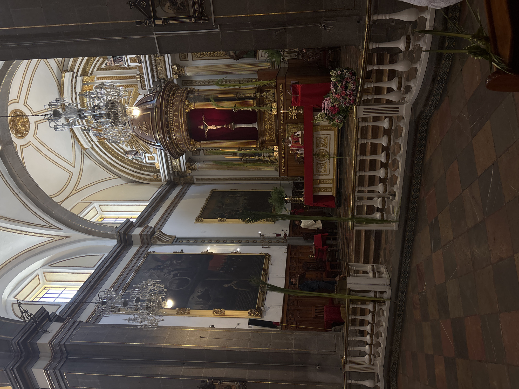 The image captures the interior of a cathedral in Puebla, Mexico. The altar features a crucifix against a red backdrop, surrounded by elaborate gold ornamentation. Crystal chandeliers hang from the vaulted ceiling. Palm branches are placed near the altar and along a white balustrade. Paintings adorn the walls. A person in white attire is visible near the altar, while a person wearing black clothing is situated in the aisle. The floor is tiled with a pattern of square and rectangular tiles. The interior is well-lit, creating a sense of grandeur.