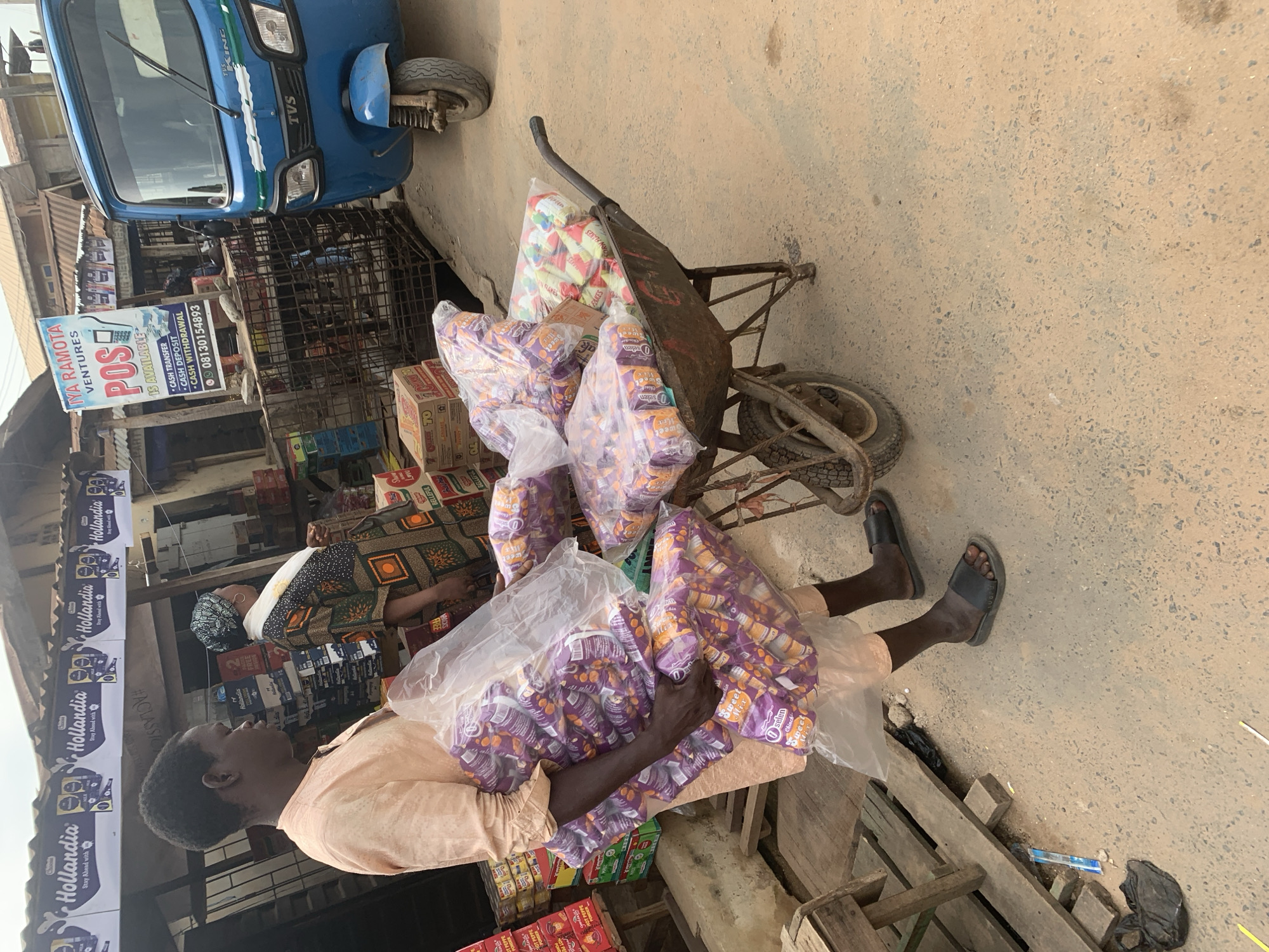 This media file captures a street scene in Shagamu, Nigeria, likely during daylight hours. The primary subjects are two people, appearing to be vendors, engaged in their work. One person, dressed in a light-colored shirt, is seen from the side, looking upwards. Another person, in the background, is partially obscured by a cart.
The scene is characterized by a cluttered marketplace environment. A blue three-wheeled vehicle, possibly a tricycle or tuk-tuk, is parked on the left side of the frame. In the center, a wheelbarrow is heavily laden with numerous plastic bags filled with packaged goods, predominantly small purple and orange packages. Several boxes of what appears to be food products are also visible. A sign that reads "IYA RAMOTA VENTURES POS IS AVAILABLE CASH TRANSFER CASH DEPOSIT CASH WITHDRAWAL 08130154893" is prominently displayed, indicating a business offering financial services. Another sign features "Hollandia" branding. The ground is unpaved and dusty, typical of a market area.
The overall impression is one of a busy, functional marketplace where goods are being transported and sold, with the individuals appearing focused on their tasks. The scene suggests a warm, perhaps humid, climate, common in Nigeria.