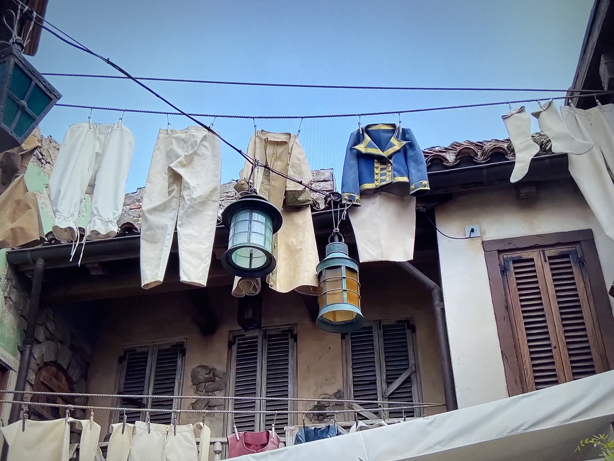 The image shows a scene from a historical-themed area, likely a theme park or outdoor museum. A clothesline is strung between buildings, and various pieces of clothing and lanterns are hanging from it. The clothing includes trousers, a tunic, a jacket with gold detailing, and stockings. The lanterns are rectangular and appear to be made of metal and glass. The buildings behind the clothesline have a rustic, Mediterranean style, with weathered walls, shutters, and tile roofs. A second clothesline is visible on a lower balcony. The location is not precisely identifiable from the image but is likely a themed environment in a location that has not been specified.
