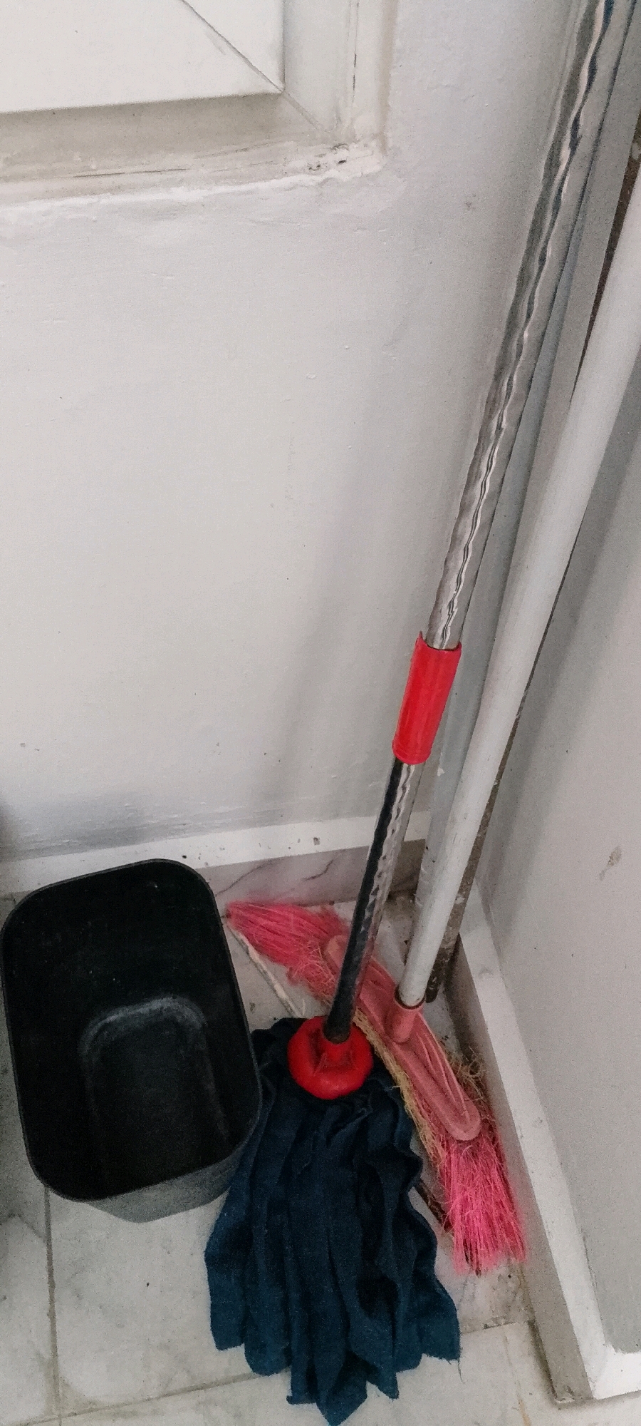 In a corner of a room, likely in City of Medan, Indonesia, a dark plastic dustpan rests on a tiled floor next to a blue microfiber mop. The mop has a red handle attachment and its strings are spread out on the floor. Behind the mop, a pink-bristled broom leans against a white wall. The wall has a white baseboard and a window frame is visible at the top of the image.