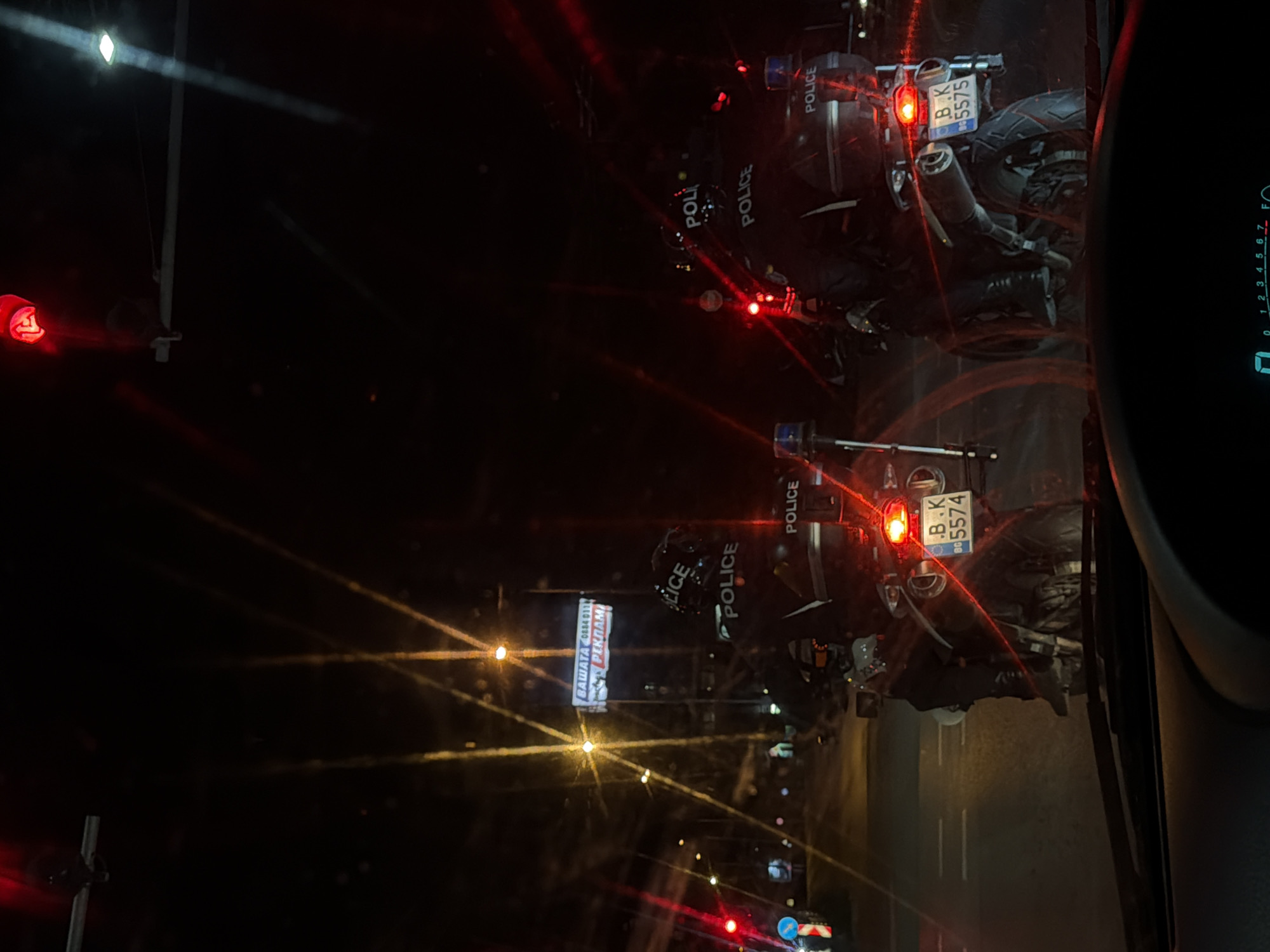 The image shows a night scene, likely taken from inside a vehicle. Two police motorcycles are visible in front, identifiable by the "POLICE" markings on their sides and the registration plates. One plate reads "B.K 5574" and the other "BK 5575". The motorcycles are emitting red light from their tail lights. Street lights and other light sources create streaks across the image due to the camera's exposure settings or condensation. A billboard with text "ВАШАТА 0884 011 РЕКЛАМ" can be seen. The location is likely in Varna, Bulgaria, given the language on the billboard and the format of the license plates. The speedometer of the vehicle taking the image is visible and shows that vehicle is not moving.