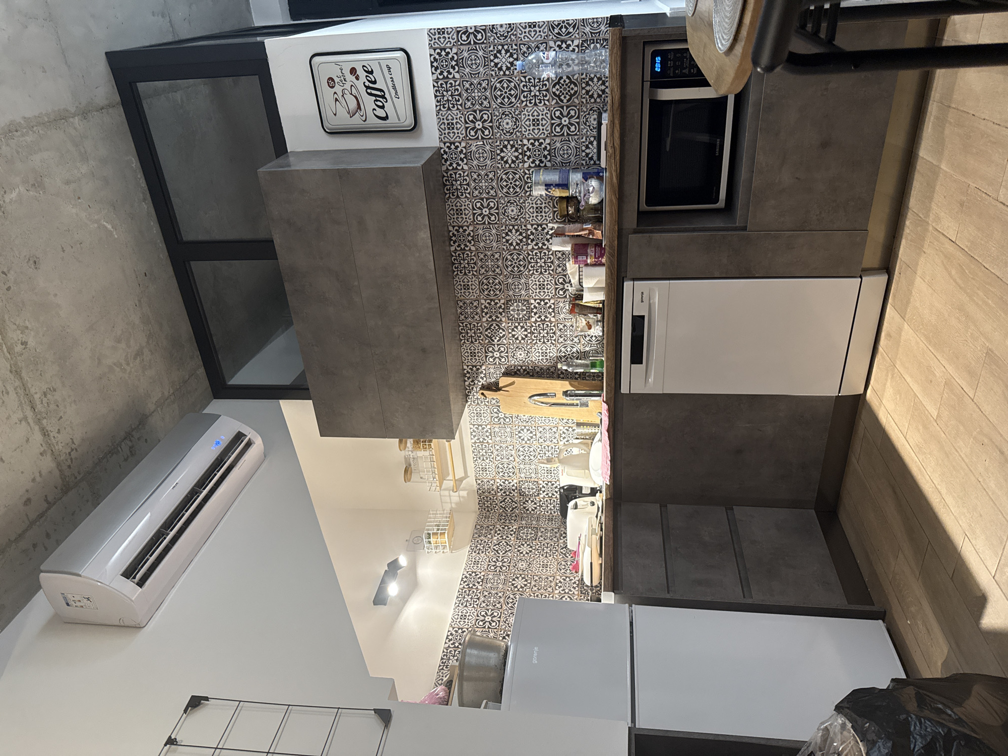 The image shows a compact kitchen area. The kitchen features a white refrigerator on the left, a white dishwasher in the center, and a black microwave oven on the right. The countertops are wooden, and the backsplash is a black and white patterned tile. Above the counter, there are several shelves with jars and containers. An air conditioning unit is mounted on the white ceiling, which also has exposed concrete elements. A framed "Coffee" sign is hanging on the wall. The floor is tiled with light-colored wooden planks. The lighting appears to be from overhead track lights.